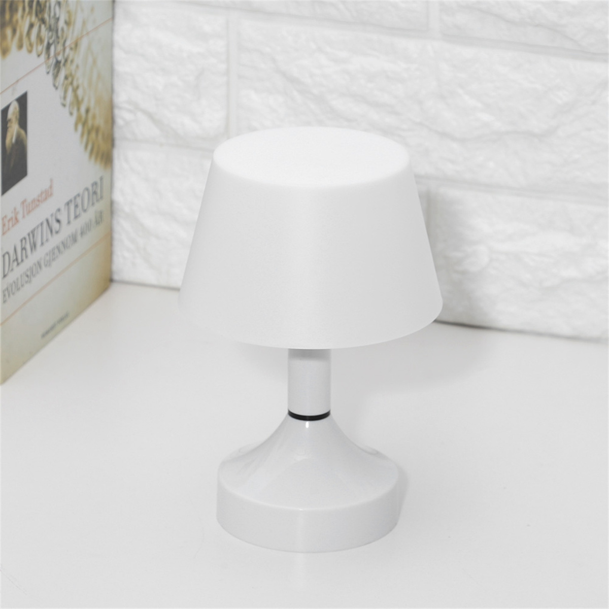 Eine weiße Lampe mit Lampenschirm steht auf einem weißen Tisch. Links von der Lampe befindet sich ein Buch.