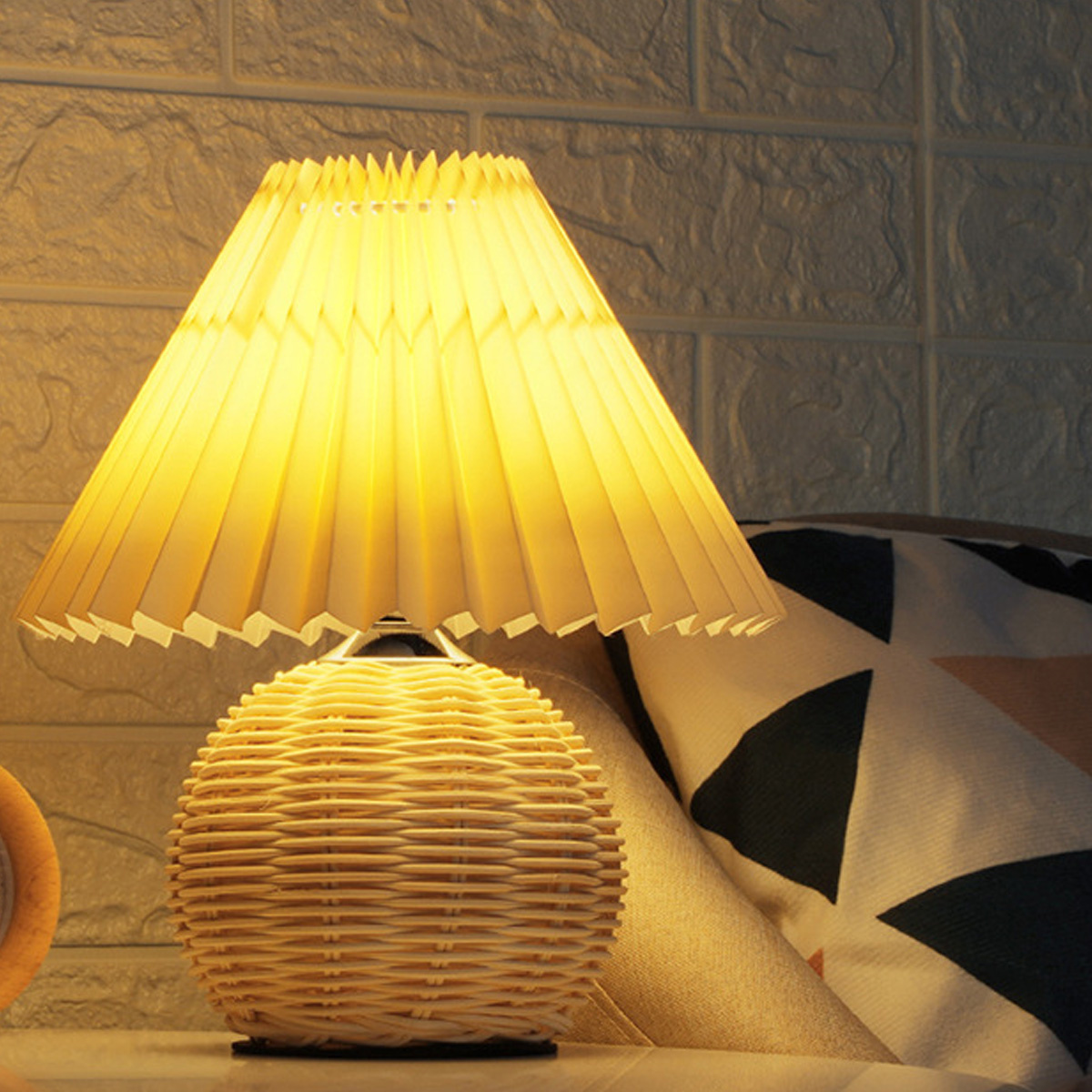 Eine Lampe mit einem Rattansockel und einem plissierten Lampenschirm, der warmgelbes Licht abgibt.