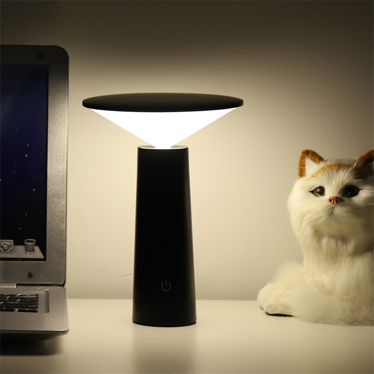 Eine schwarze moderne Lampe steht auf einem weißen Tisch. Ein Laptop und eine Katzenfigur sind auch vorhanden.