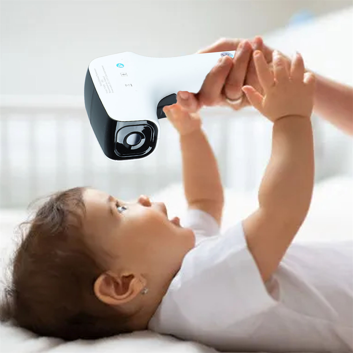 Ein Baby blickt auf ein Thermometer, das von einer Hand gehalten wird. Das Baby liegt auf einem weißen Bett.