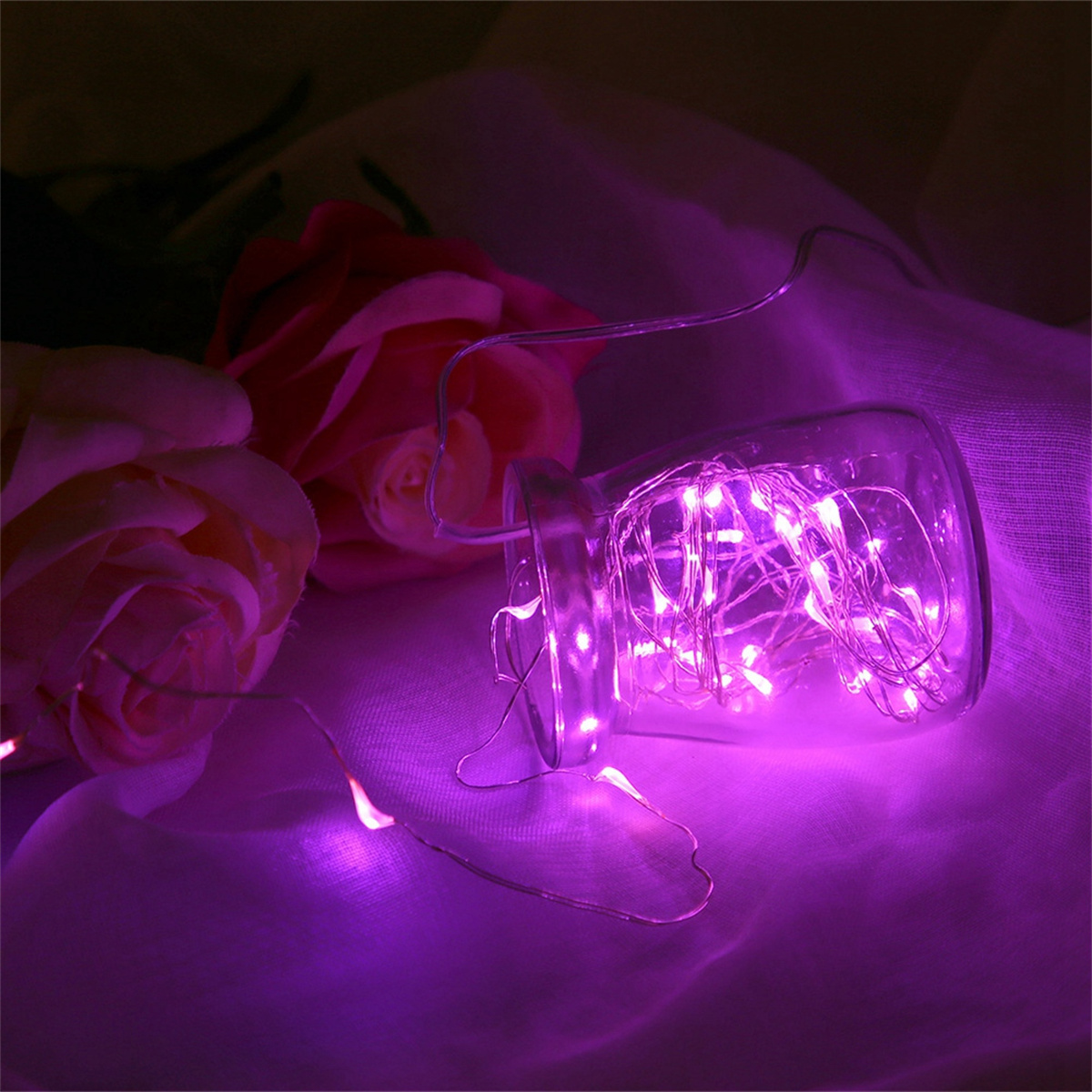 Beleuchtete Lichterketten in einem Glas neben Rosen. Lila leuchten. Weißer Stoffhintergrund.
