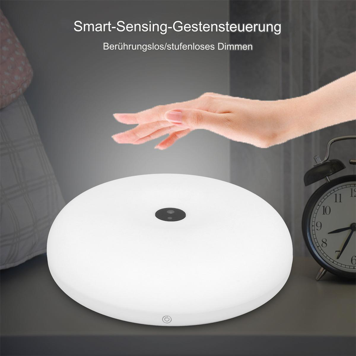 Eine weiße, kreisförmige Lampe mit einer Hand darüber. Im Hintergrund ein Nachttisch.