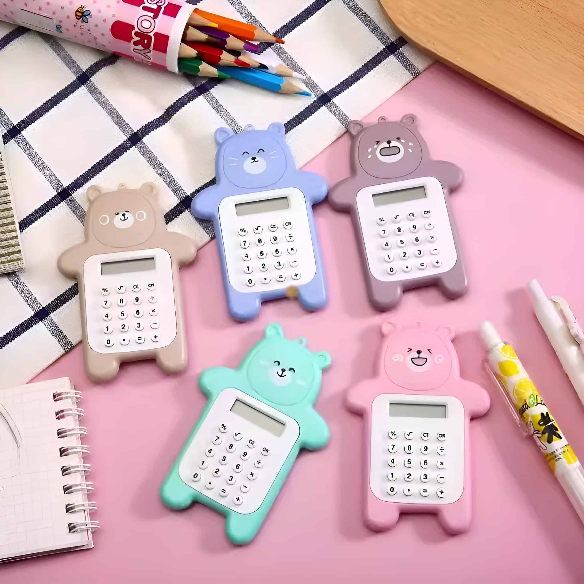 Bärenförmige Taschenrechner in verschiedenen Farben: Beige, Blau, Lila, Grün und Rosa. Buntstifte und Stift daneben.