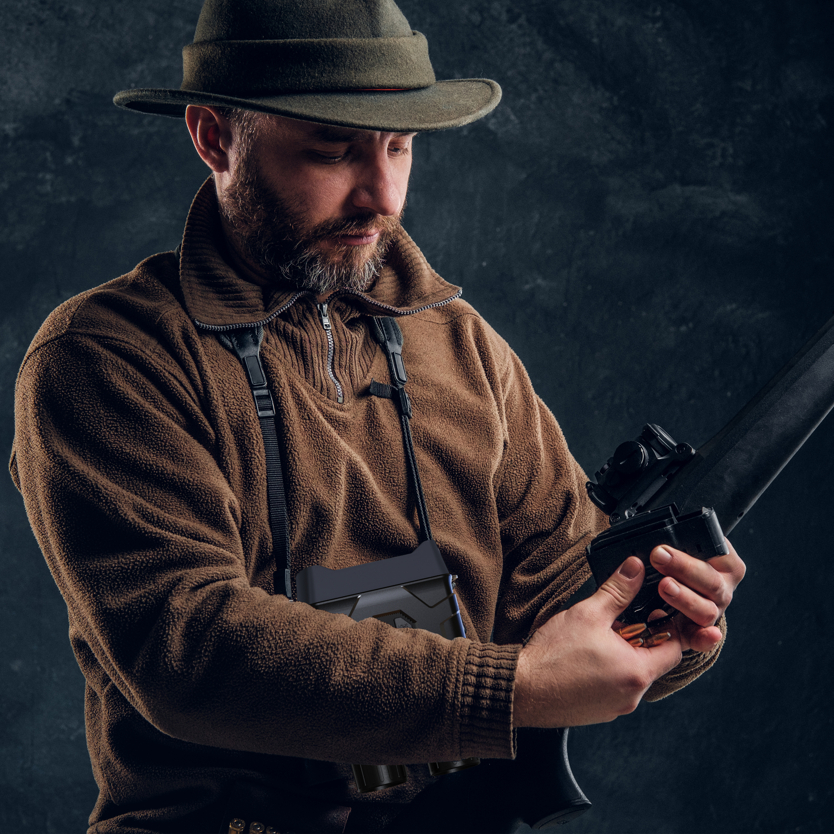 Mężczyzna w stroju myśliwskim trzymający karabin, z kapeluszem i lornetką, ciemne tło.