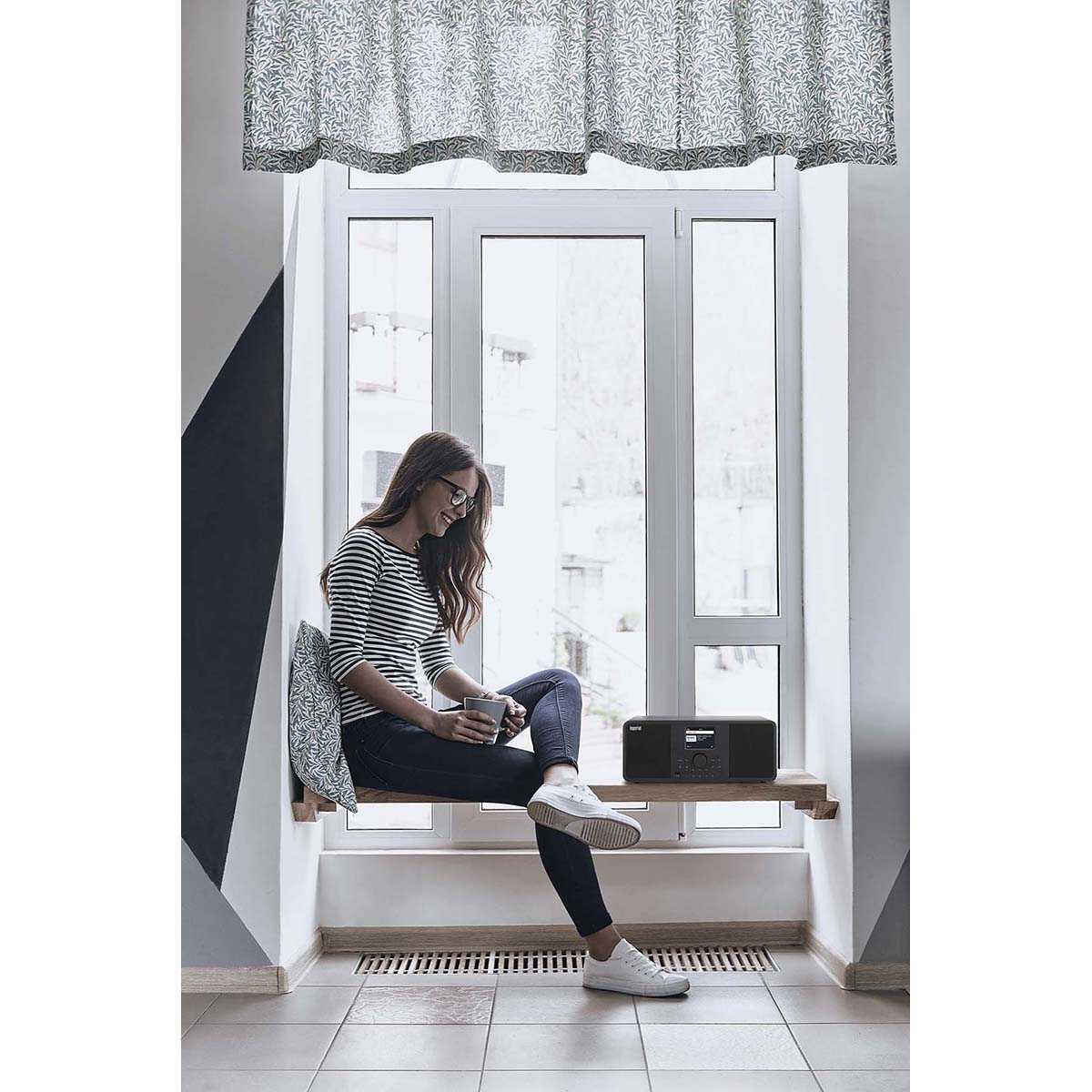 Kobieta siedząca przy oknie z radiem na półce i wzorzystą zasłoną.