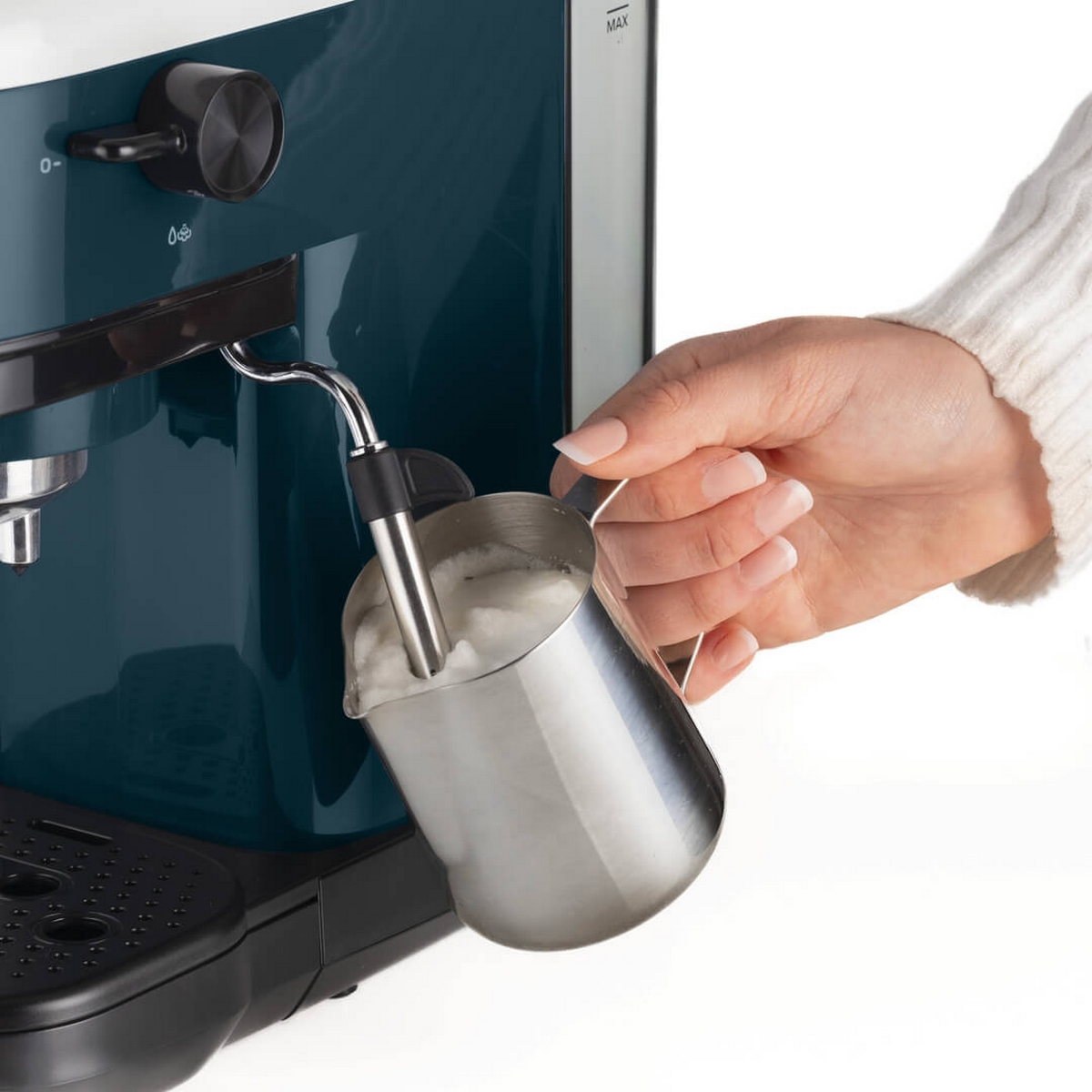 Ręka wlewa spienione mleko do metalowego dzbanka. Po lewej stronie znajduje się turkusowy ekspres do kawy. Maszyna stoi na blacie.