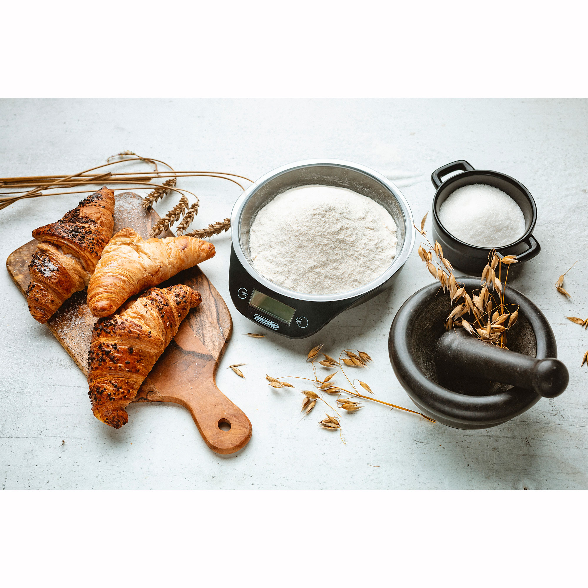 Escena de horneado con cruasanes, balanza con harina, azucarero y mortero con avena.
