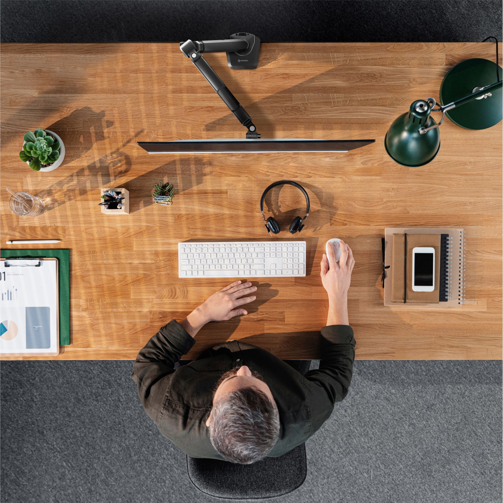 Widok z góry na drewniane biurko. Mężczyzna siedzi. Zawiera monitor, klawiaturę, słuchawki i lampę.