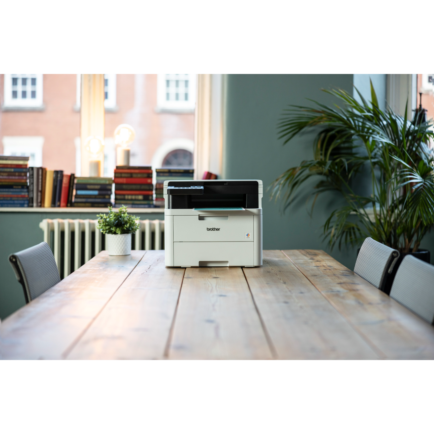 Drukarka Brother na drewnianym stole, z oknem i rośliną w tle. Pokój ma zielone ściany.