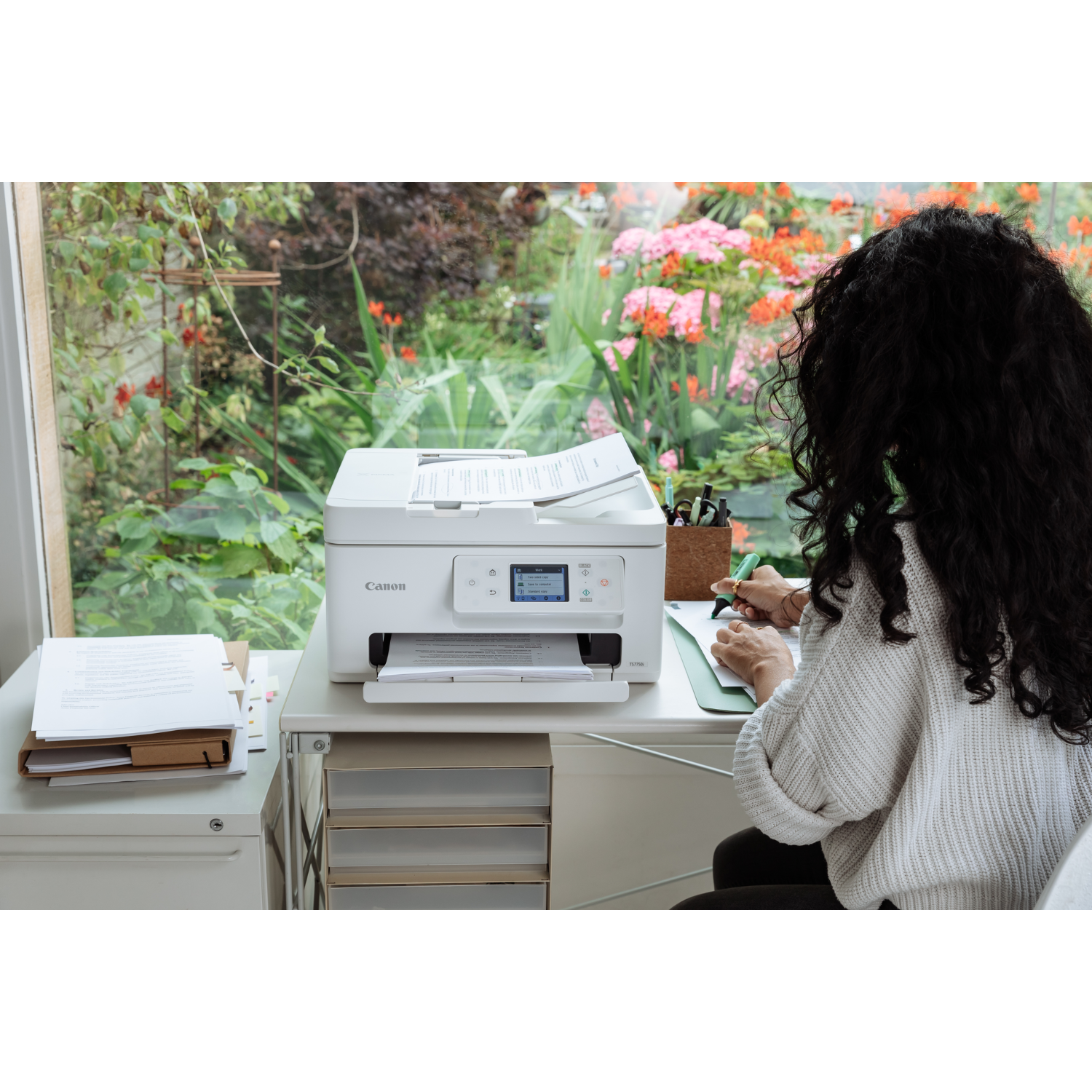 Kobieta siedzi przy biurku, używa drukarki i pisze na notatniku. W tle okno z ogrodem.