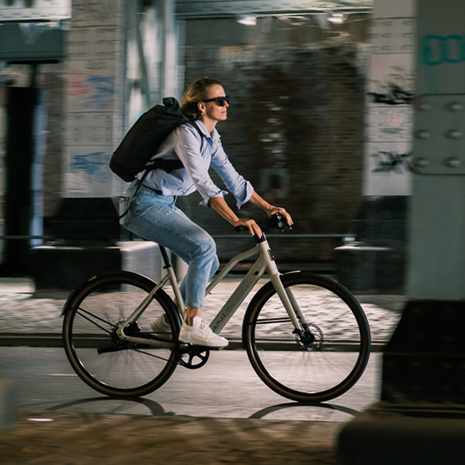 Frau auf einem Fahrrad, fährt auf einer Stadtstraße, mit Rucksack und Sonnenbrille, bei Nacht.