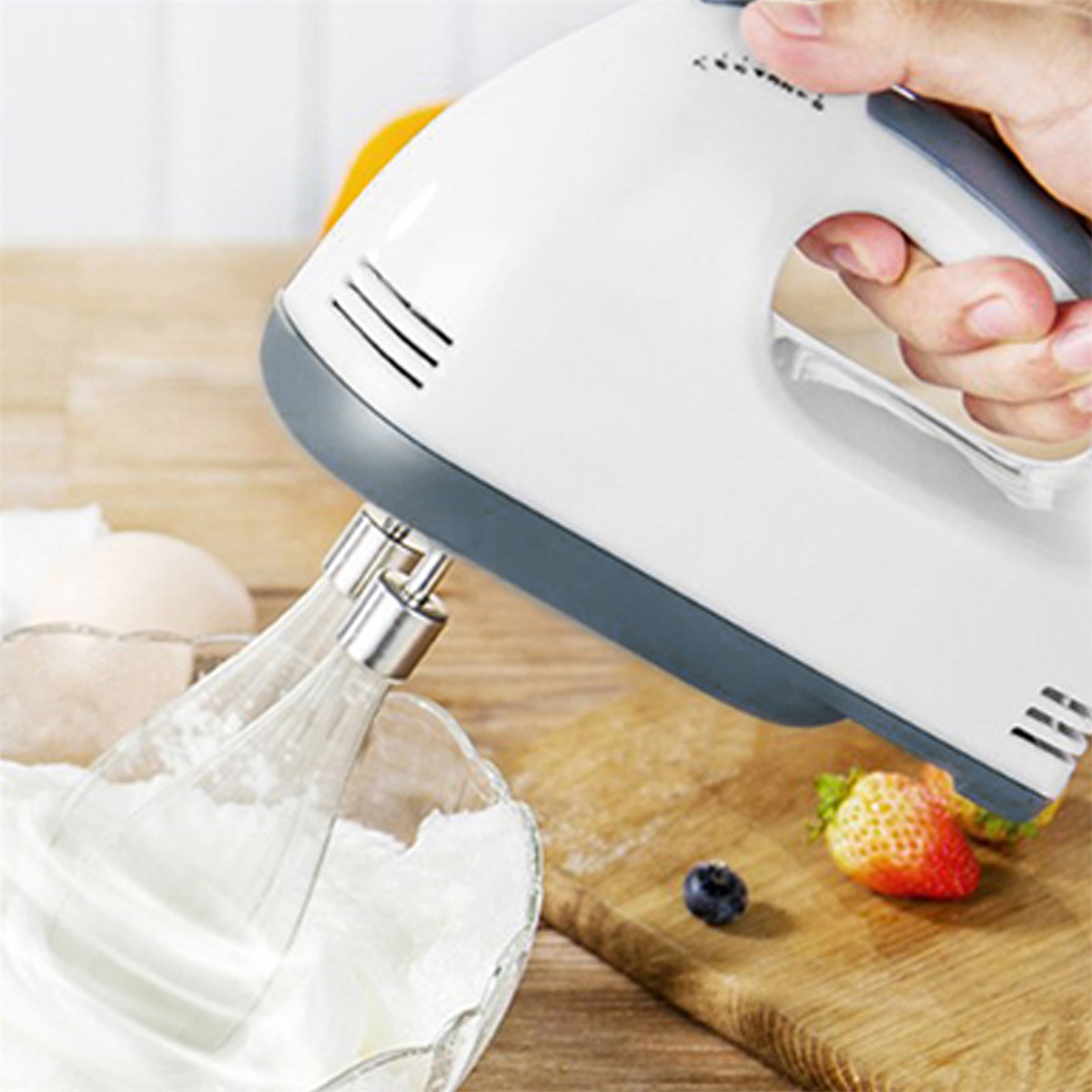Ein weißer Handmixer verrührt Zutaten in einer Glasschüssel. Erdbeeren und Blaubeeren liegen auf einem hölzernen Schneidebrett.