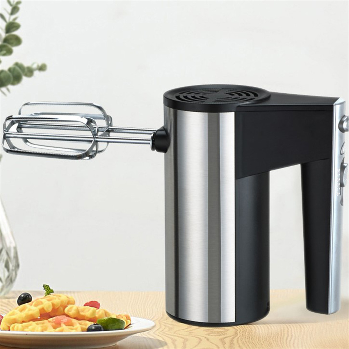 Ein silberner und schwarzer Handmixer. Ein Teller mit Waffeln und Beeren steht in der Nähe des Mixers.