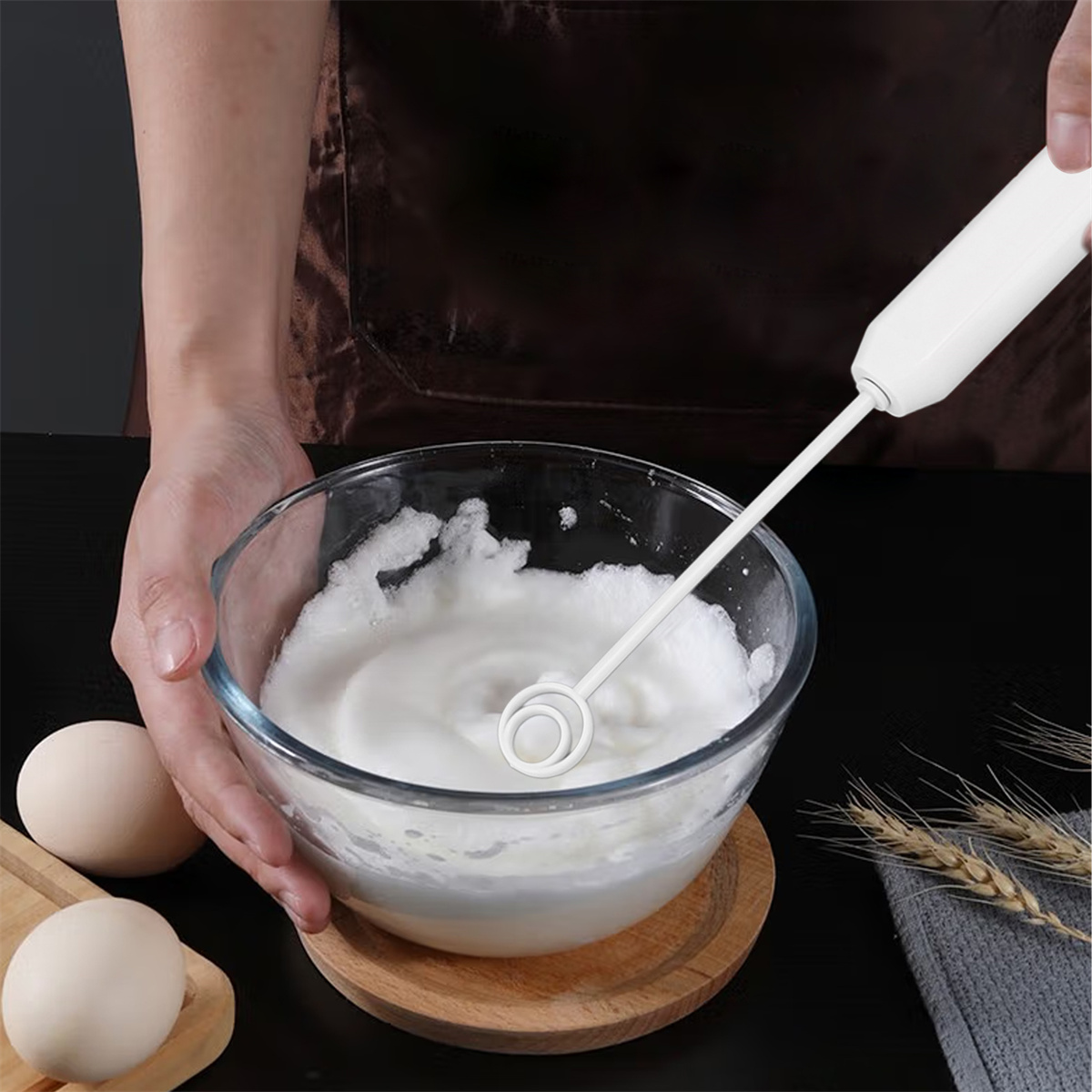 Hände, die Eischnee in einer Glasschüssel mit einem elektrischen Mixer aufschlagen, auf einem Holztablett mit Eiern.