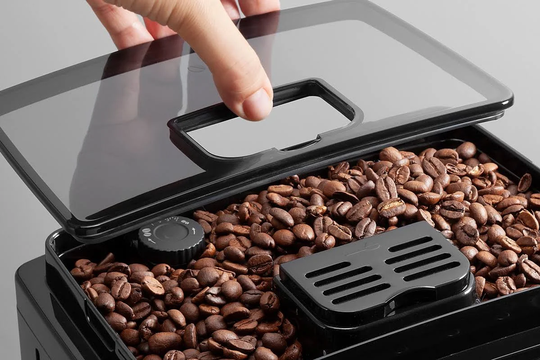 Ręka podnosząca przezroczystą pokrywę z czarnego pojemnika na ziarna kawy, ukazująca brązowe ziarna w środku.