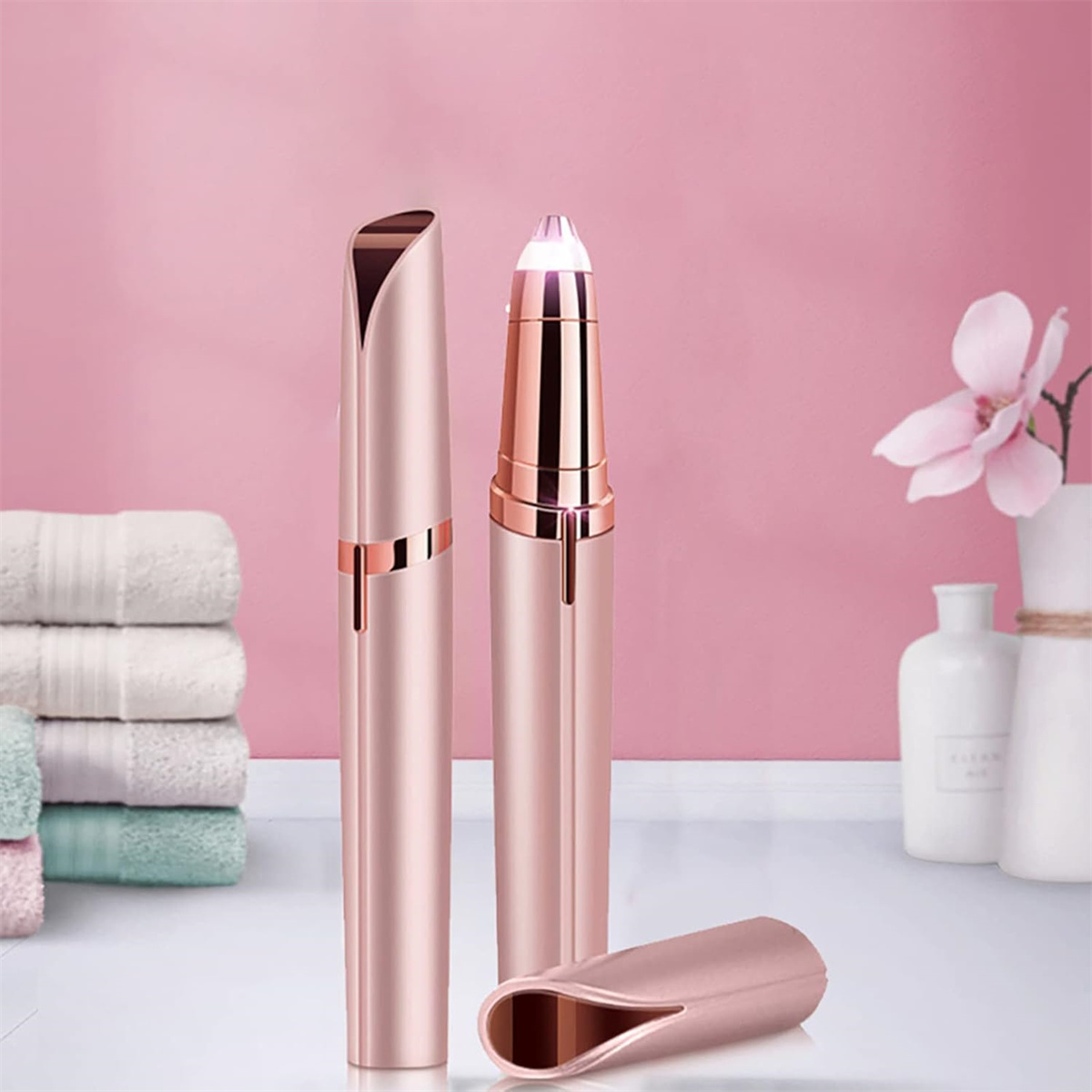 Roségoldene Gesichtshaarentferner. Zwei aufrecht, eine Kappe entfernt. Pinker Hintergrund mit Handtüchern und einer Blume.