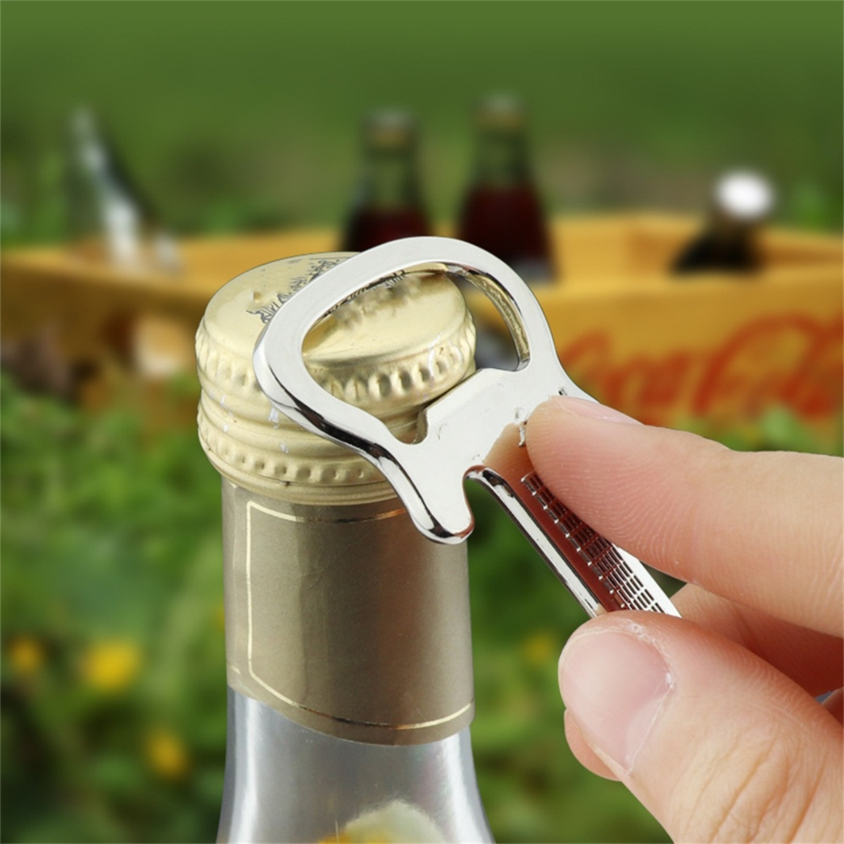 Eine Hand benutzt einen Flaschenöffner auf einer Flasche. Grünes Gras und Flaschen im Hintergrund. Glänzender Öffner.