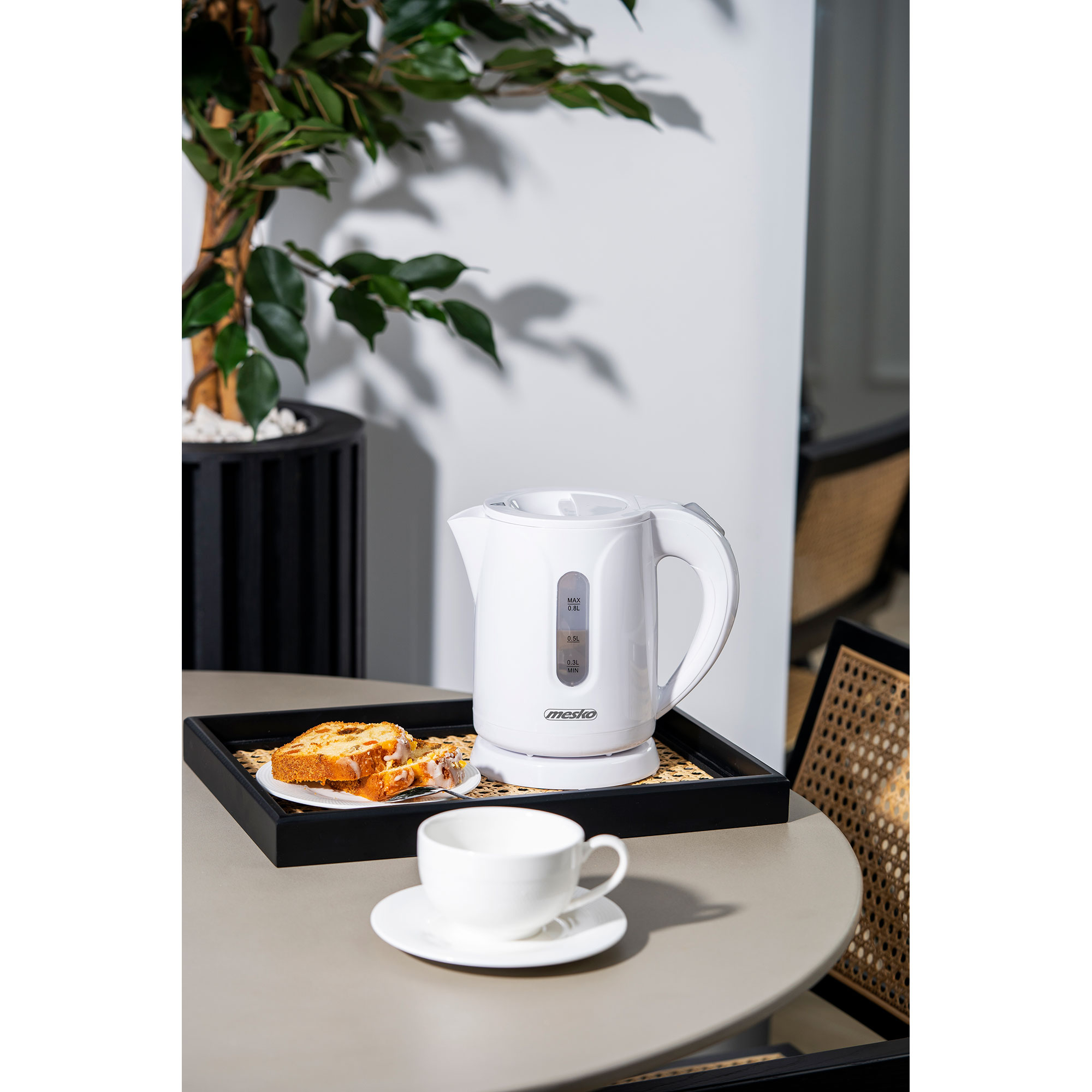 Una tetera eléctrica blanca en una bandeja con taza y pastel, cerca de una planta.