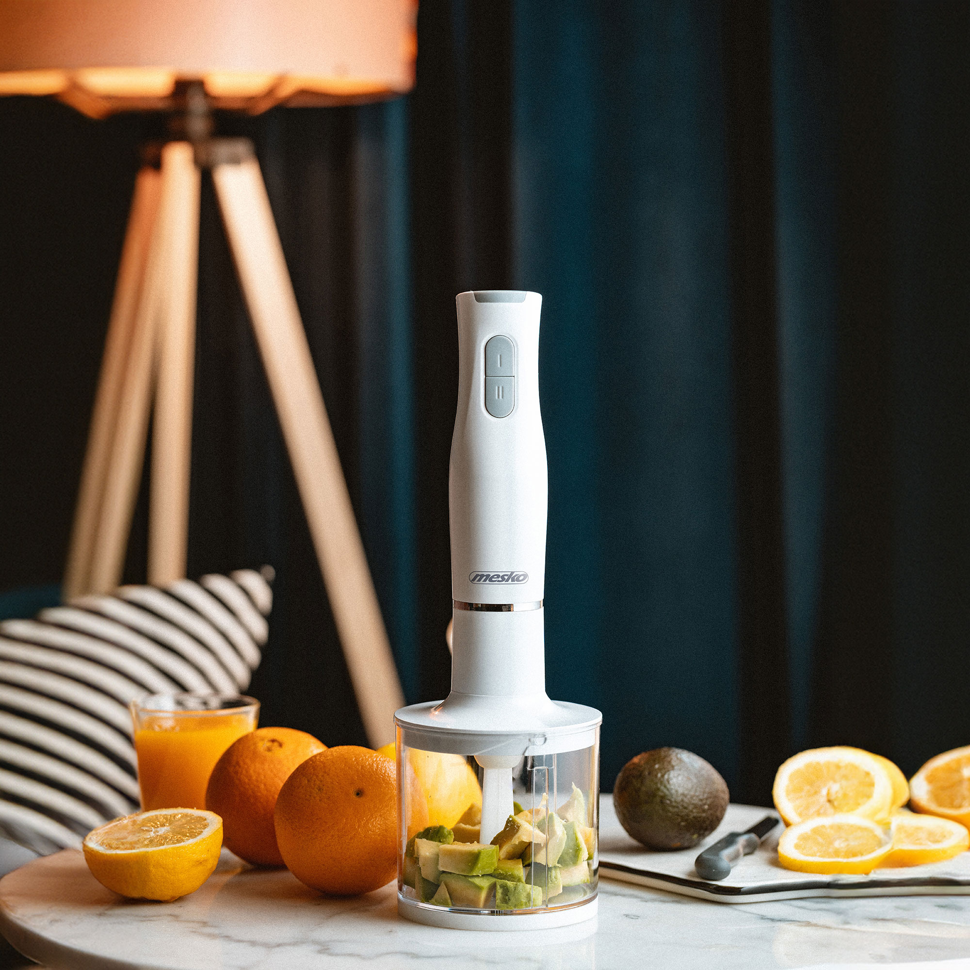 Weißer Stabmixer mit Zutaten im Glas und Orangen auf dem Tisch.