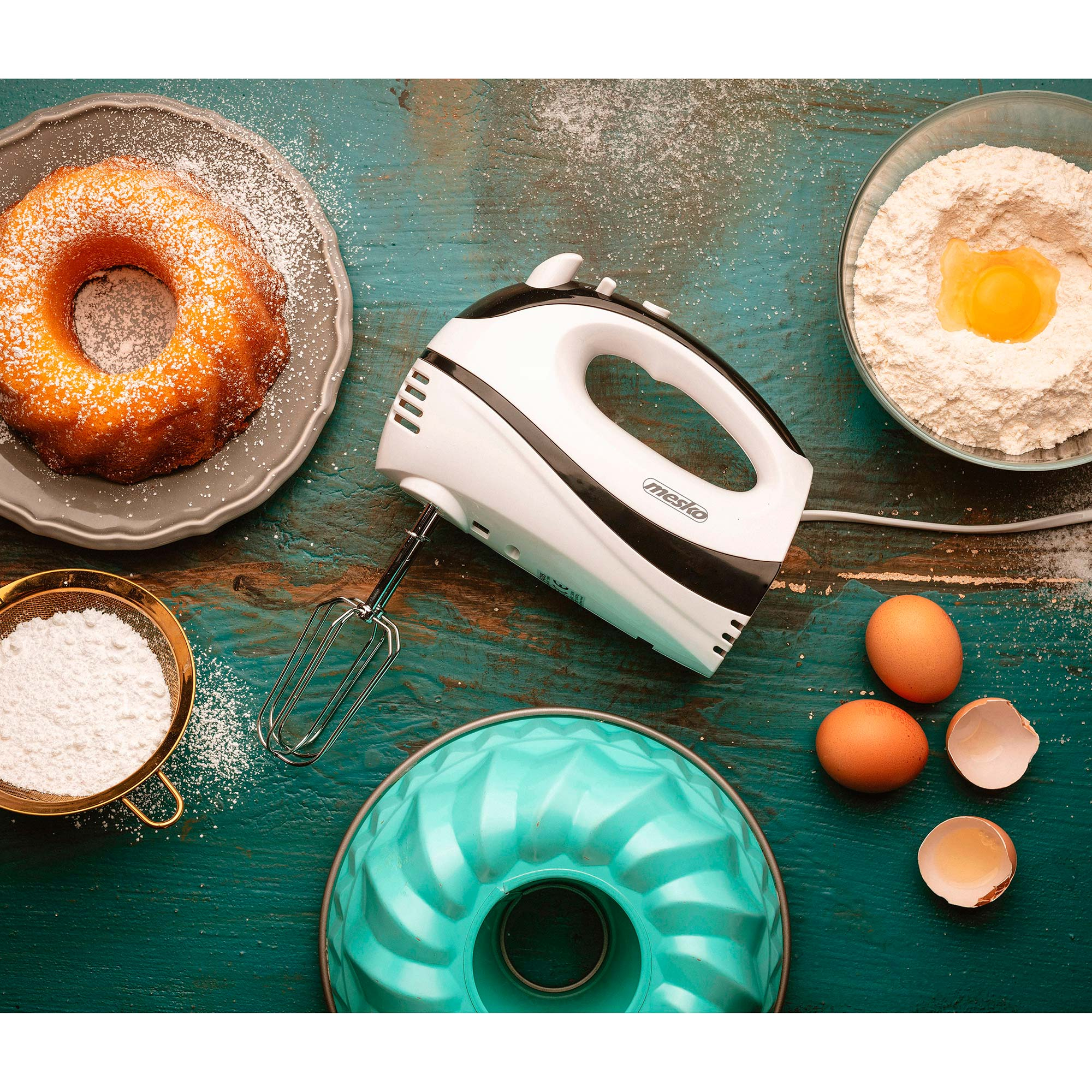 Escena de repostería con pastel, batidora, ingredientes y molde azul.