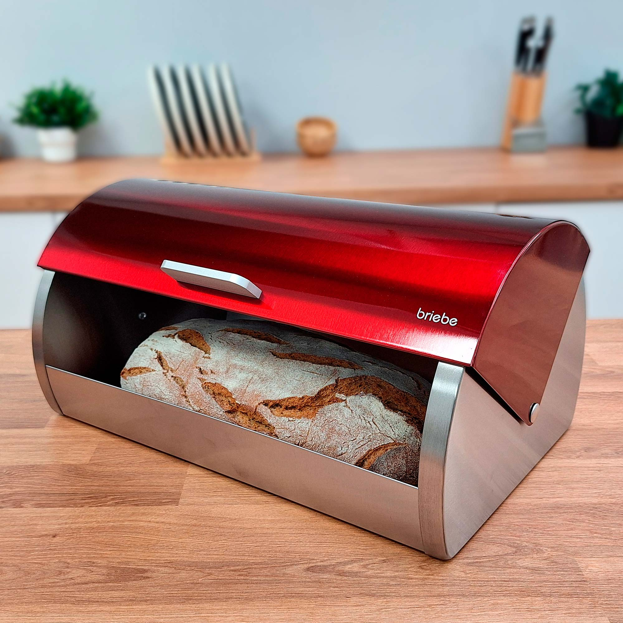 Ein roter Brotbehälter mit einem Brotlaib darin, auf einer Holzoberfläche. Der Deckel ist leicht geöffnet.