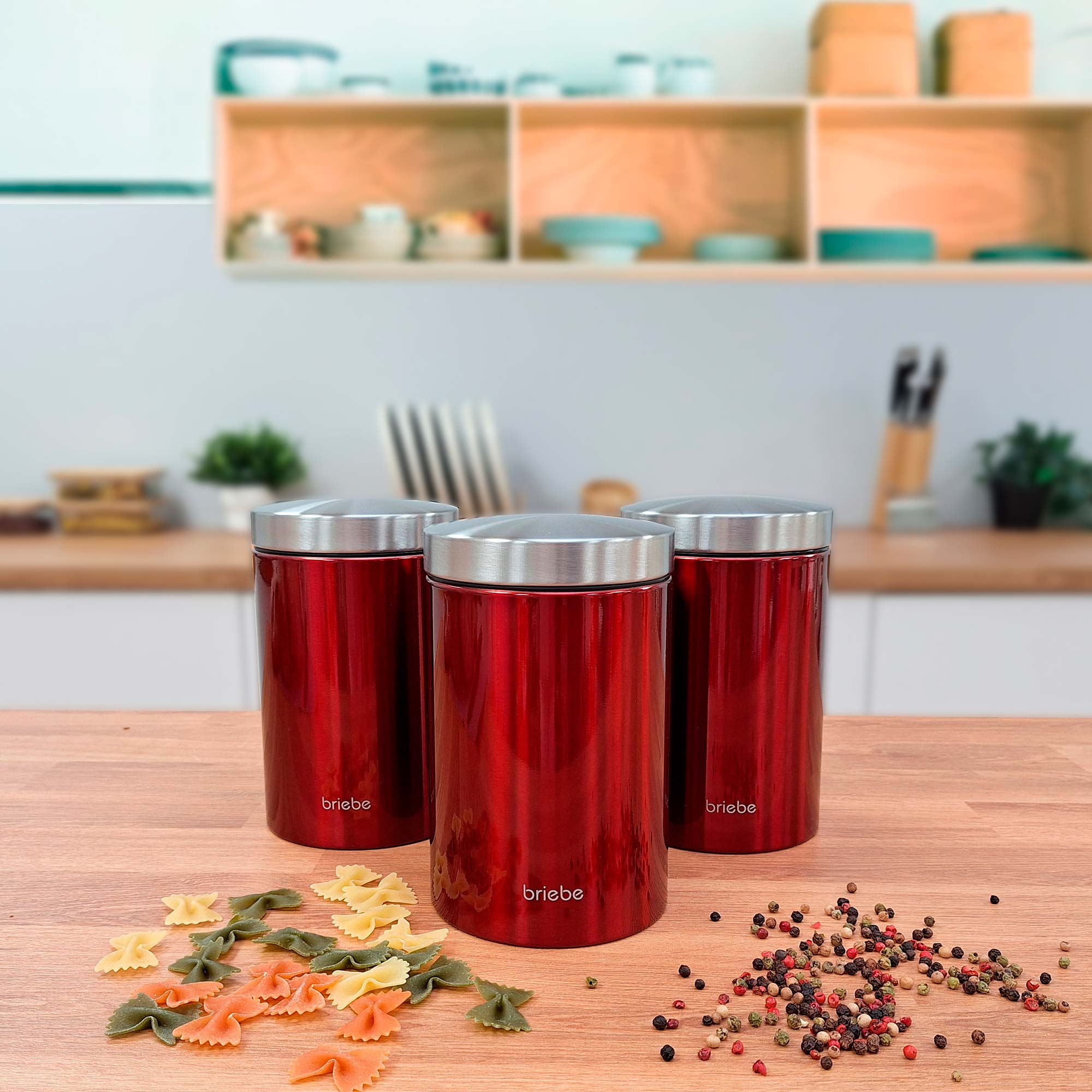 Drei rote Kanister mit silbernen Deckeln auf einer Holzoberfläche mit Pasta und Gewürzen.