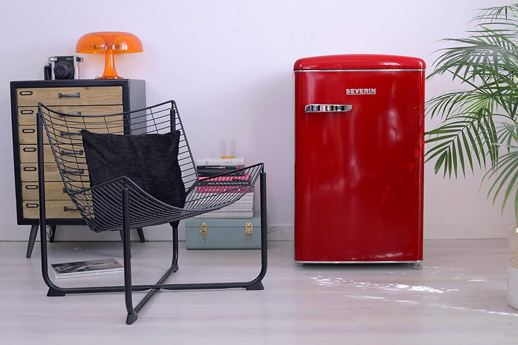 Roter Retro-Kühlschrank. Schwarzer Metallstuhl. Sideboard mit Orangenlampe und Pflanzen. Weißer Hintergrund.