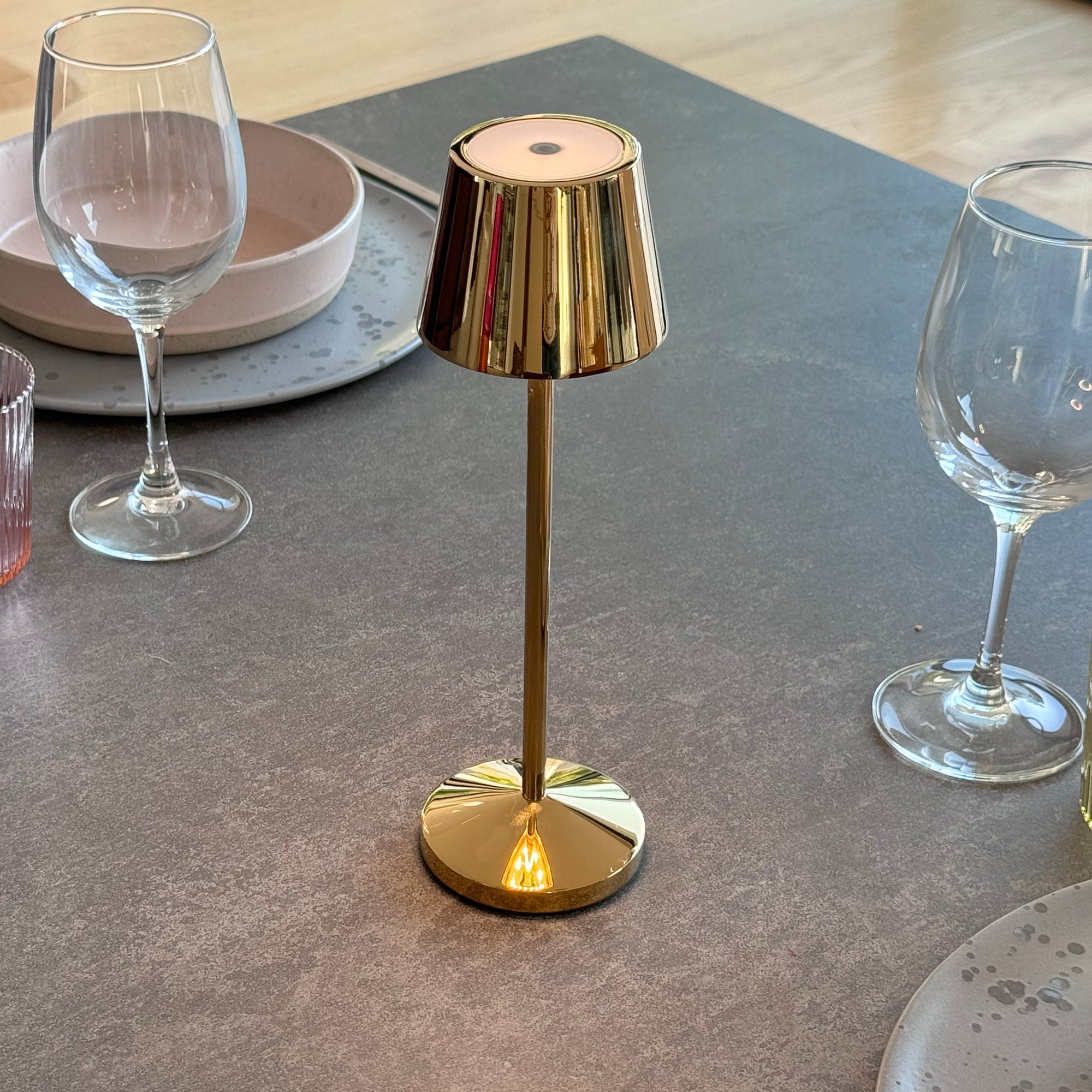 Eine goldene Tischlampe mit Schirm auf einem grauen Tisch. Weingläser und eine Schüssel sind im Hintergrund.