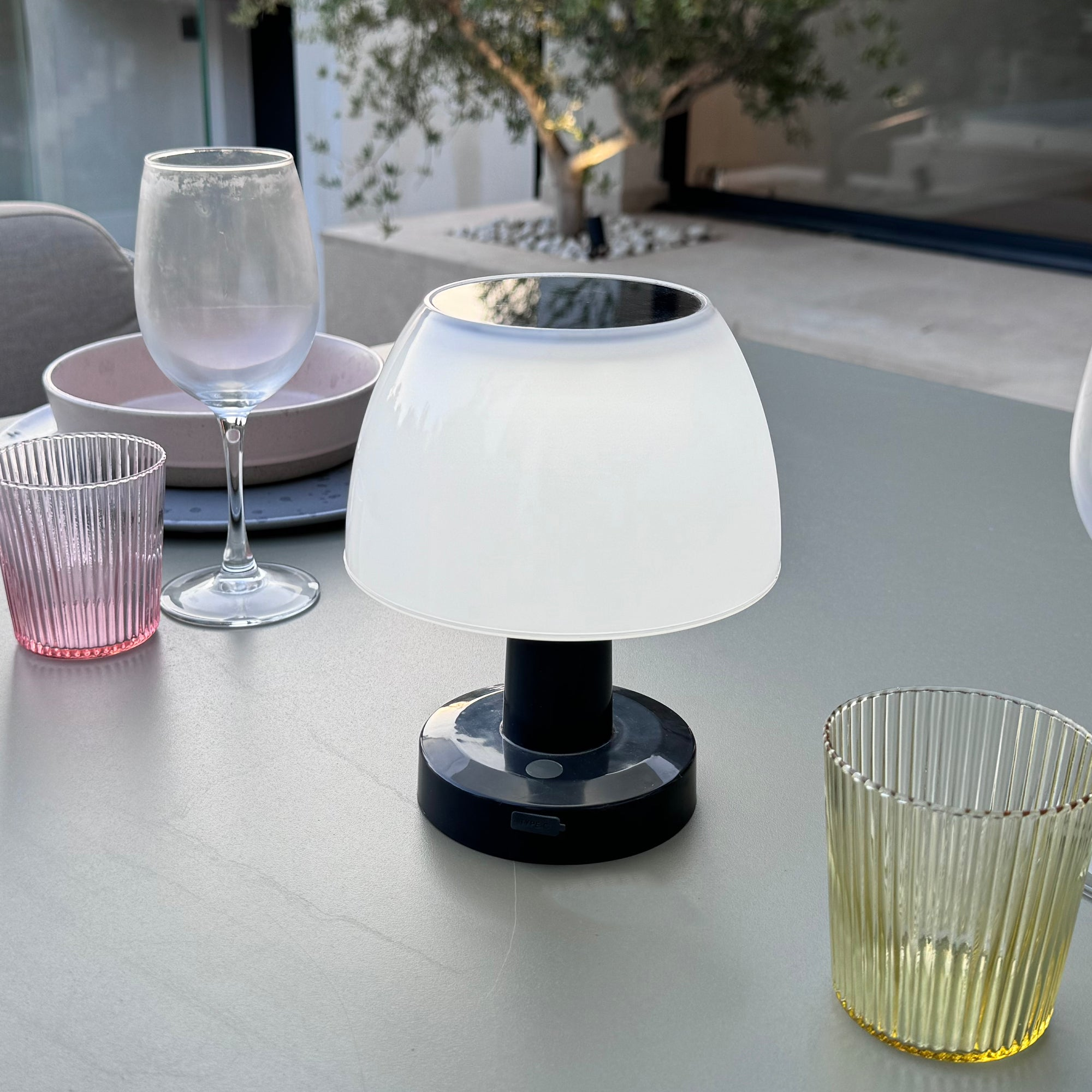 Szene im Freien: Tisch, Lampe, Gläser und Hintergrundbaum mit Sonnenlicht.