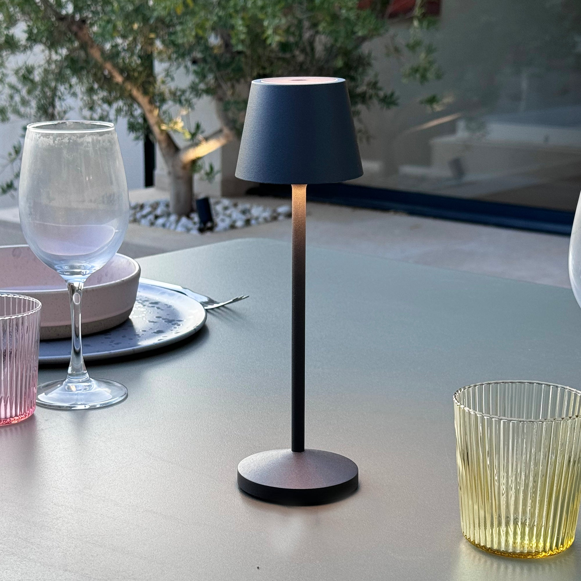 Eine schwarze Lampe auf einem Tisch mit dunklem Schirm. Im Hintergrund Gläser und Teller.