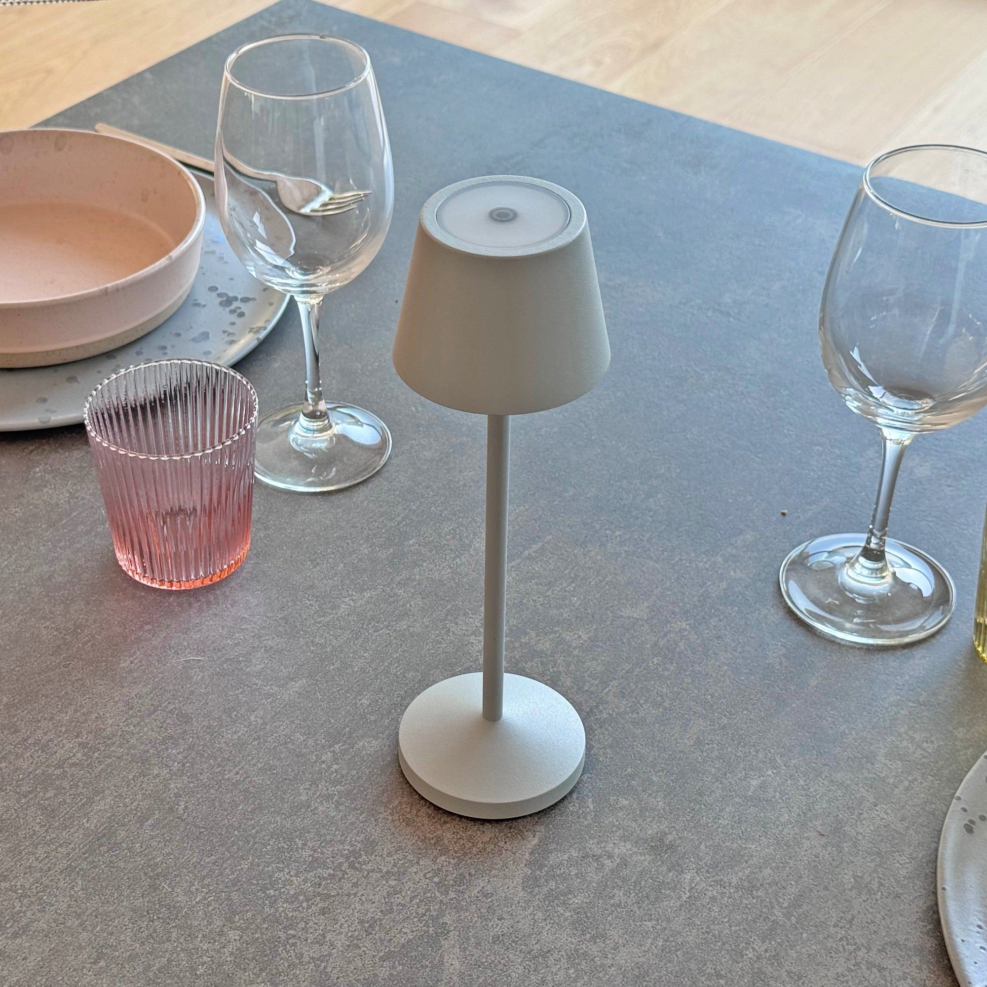 Eine weiße Tischlampe mit grauem Lampenschirm, einem rosa Glas und einem Weinglas auf einem grauen Tisch.