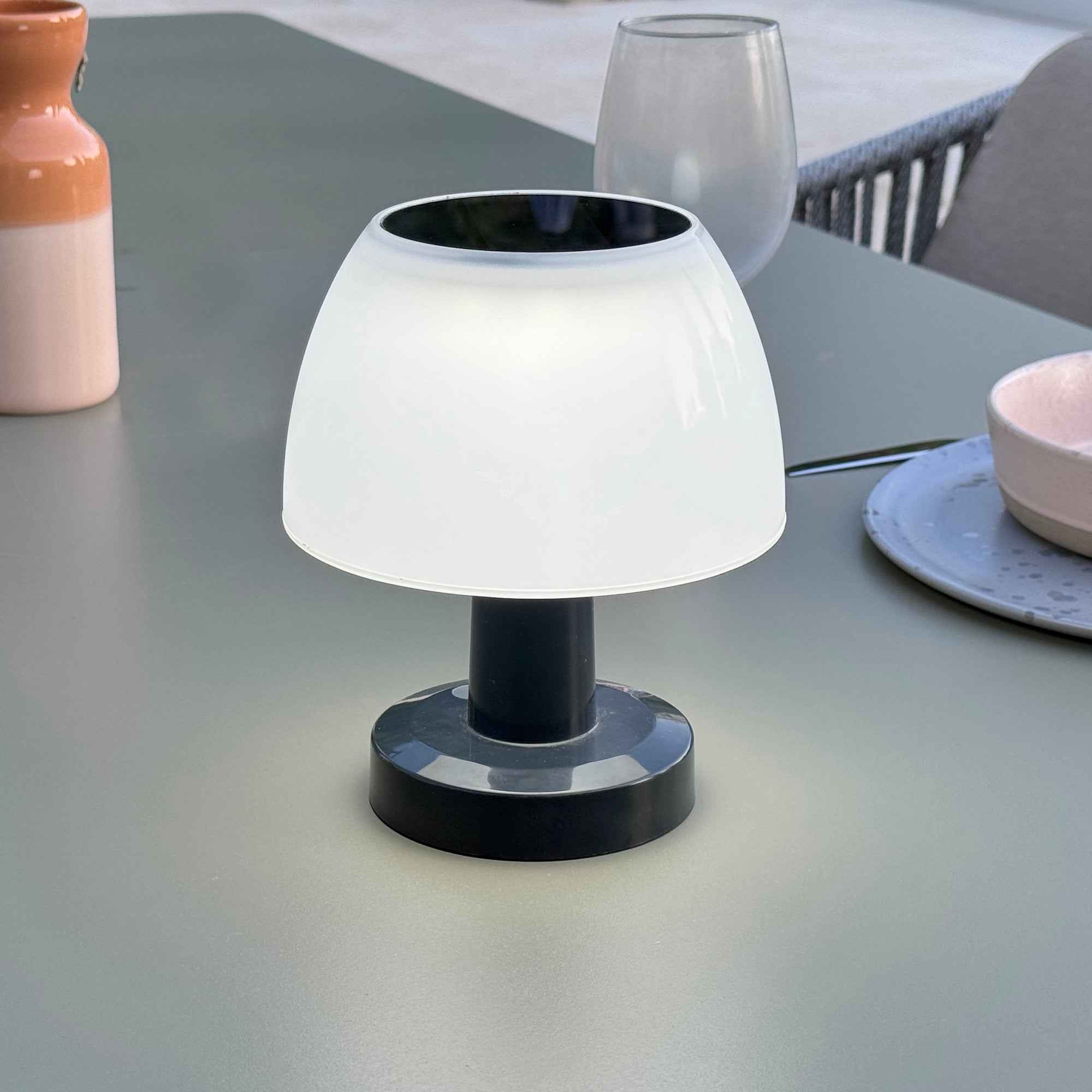 Eine Tischlampe mit weißem Schirm und schwarzem Fuß. Sie steht draußen auf einem Tisch in der Nähe einer Vase und eines Tellers.