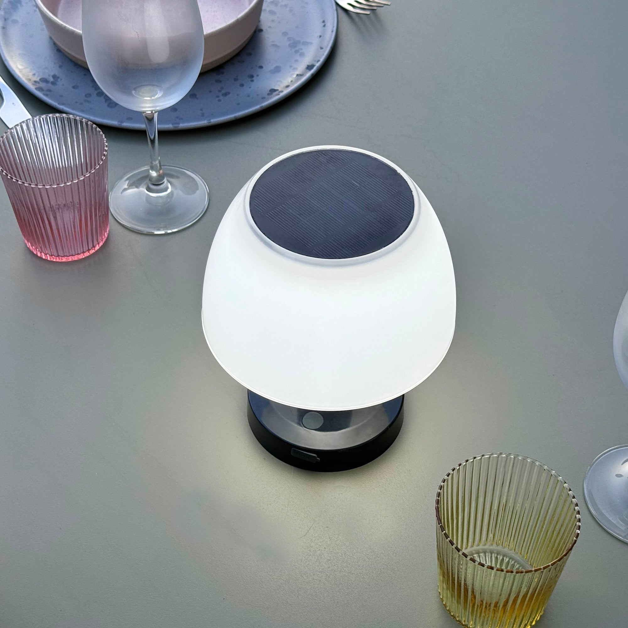 Weiße Tischlampe mit dunklem Sockel. Sie steht auf einem Tisch mit Gläsern und Tellern.