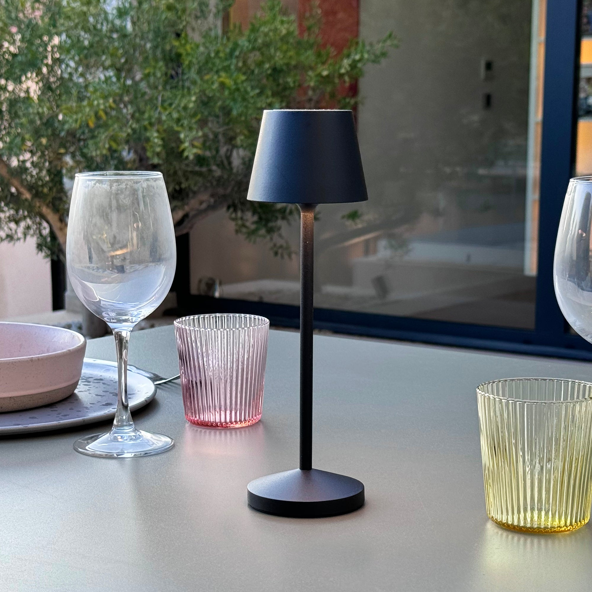 Schwarze Tischlampe auf einem Tisch mit Gläsern und anderen Gegenständen. Pflanzen im Hintergrund.