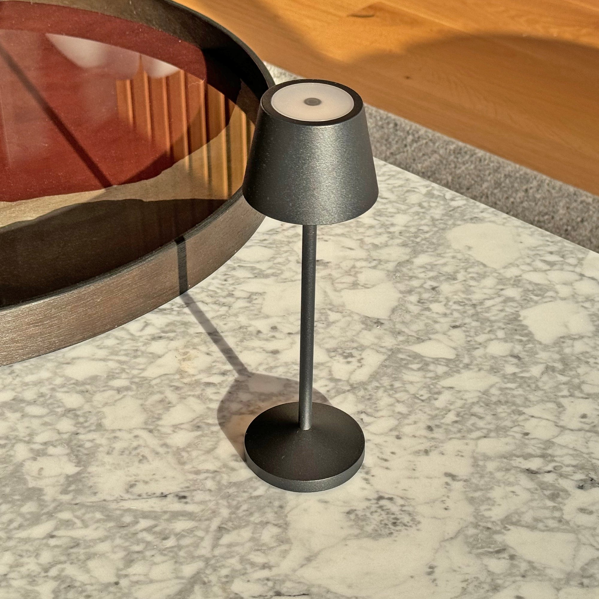 Eine schwarze Lampe auf einem Marmortisch, mit einem Glastablett und Holzboden im Hintergrund.