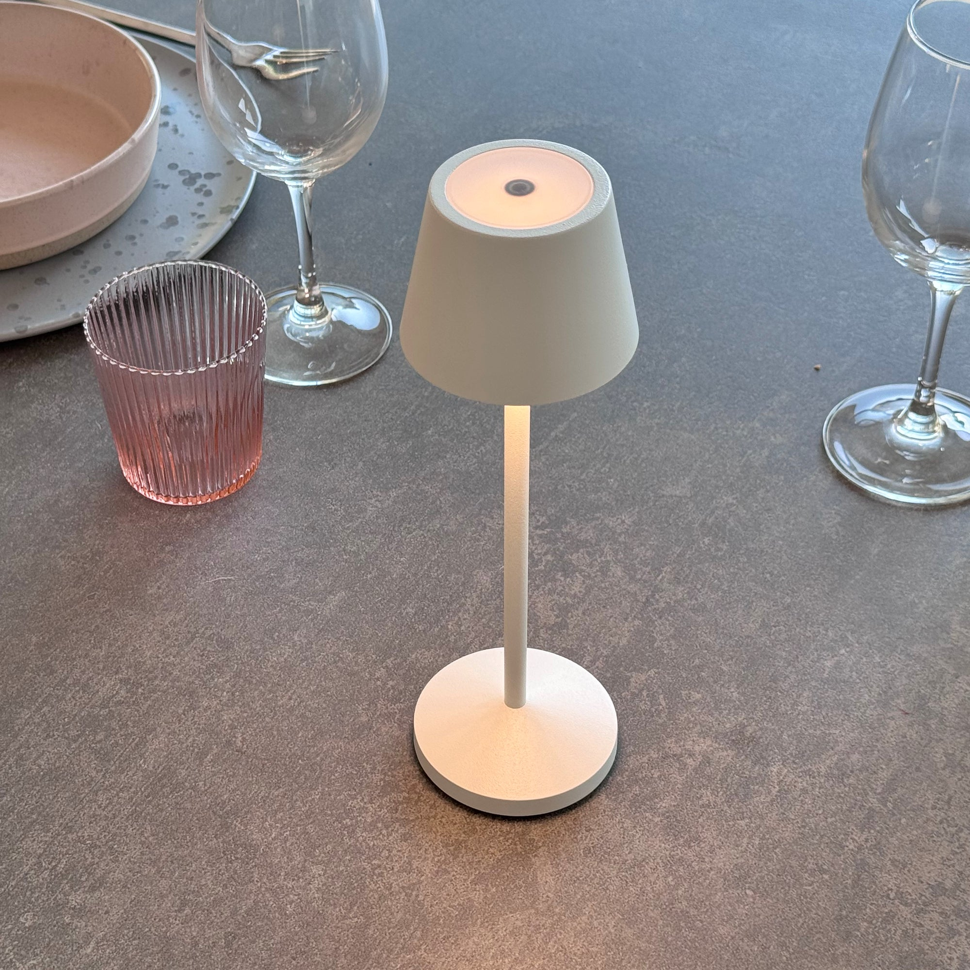 Eine weiße Lampe mit rundem Fuß und konischem Lampenschirm steht auf einem grauen Tisch. Gläser umgeben sie.