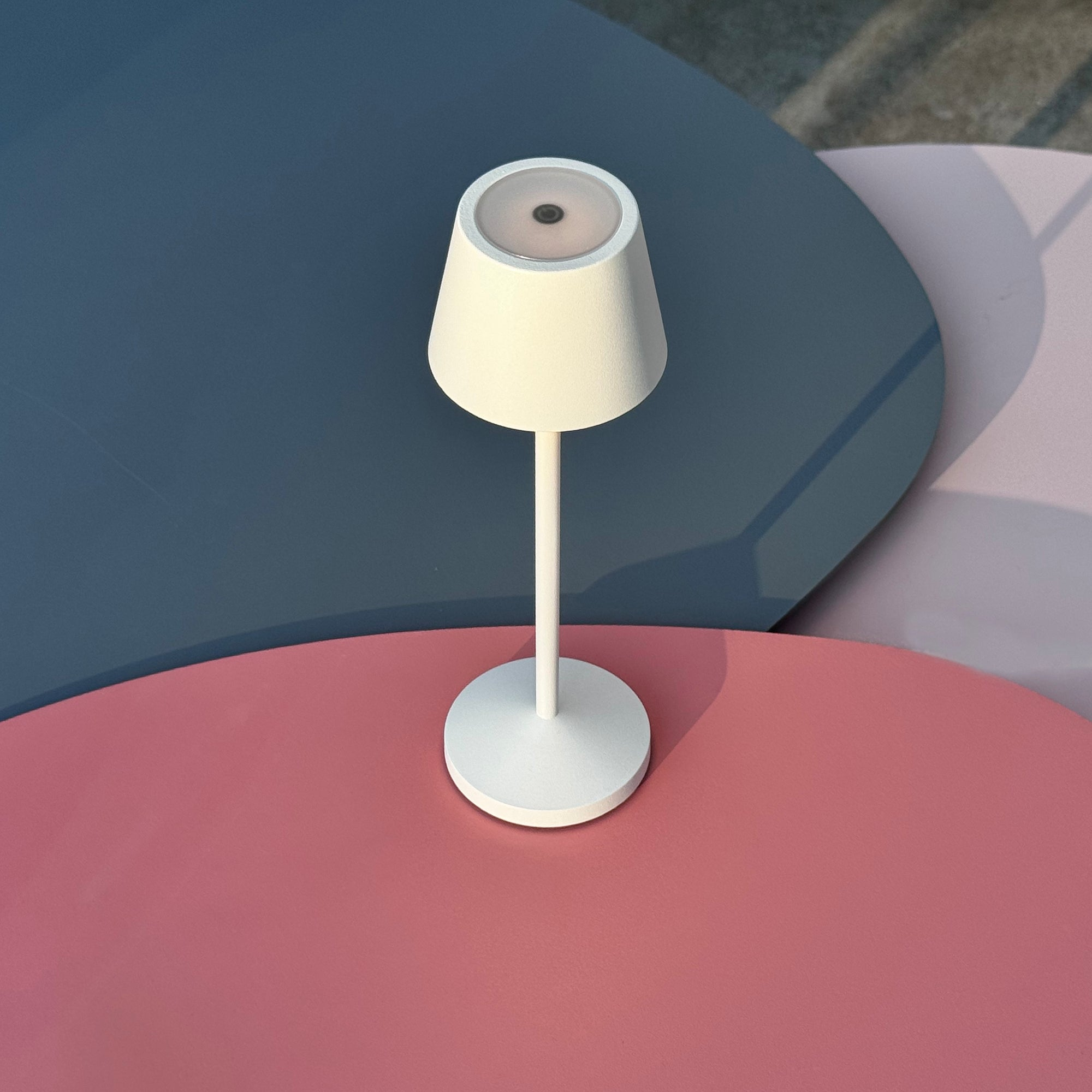 Weiße Lampe auf einem weißen Ständer, auf einer rosa, blauen und grauen Oberfläche.