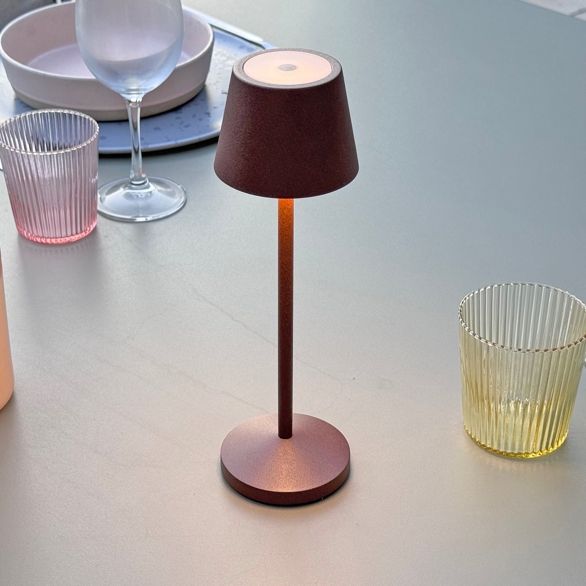 Eine braune Nachttischlampe mit Lampenschirm auf einem Tisch mit Gläsern und einem Teller.