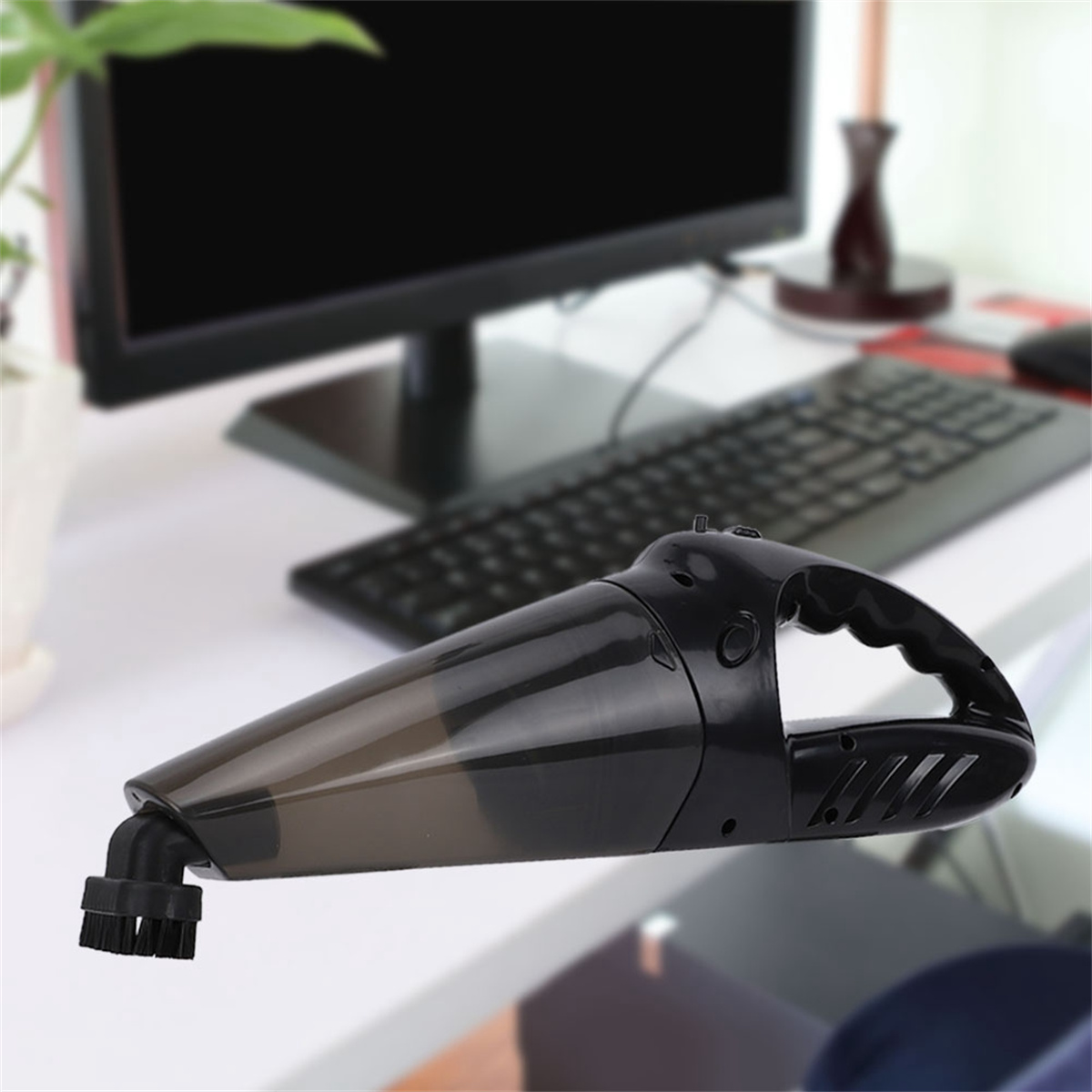 Schwarzer Handstaubsauger auf weißem Schreibtisch. Computer, Tastatur und Topfpflanze im Hintergrund.