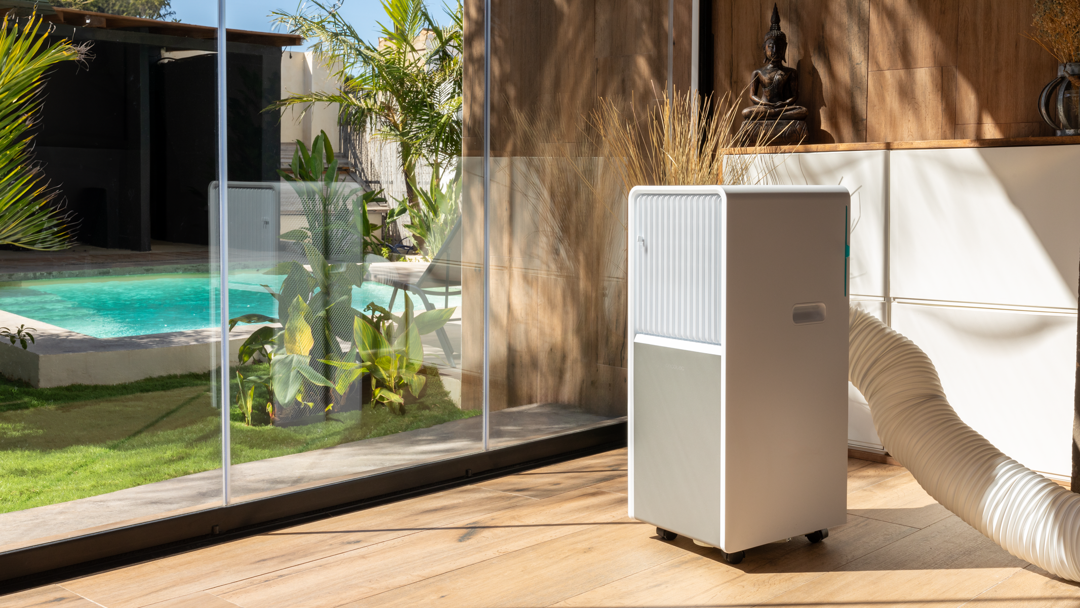 Een wit en grijs draagbaar airconditioner met een slang, geplaatst bij glazen deuren met uitzicht op een zwembad.