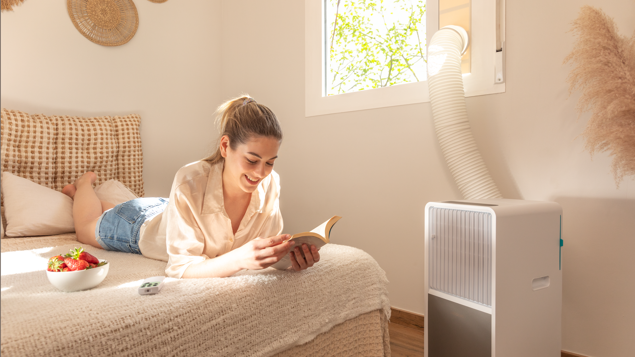 Vrouw leest boek op bed. Aardbeien in kom. Draagbare airco op de achtergrond.