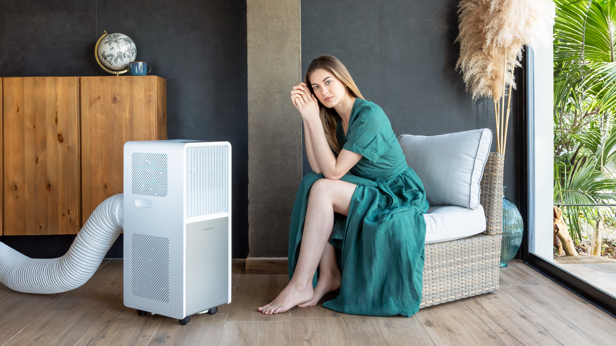 Vrouw in groene jurk zit bij een witte draagbare airconditioner. Interieur met houten kast en planten.