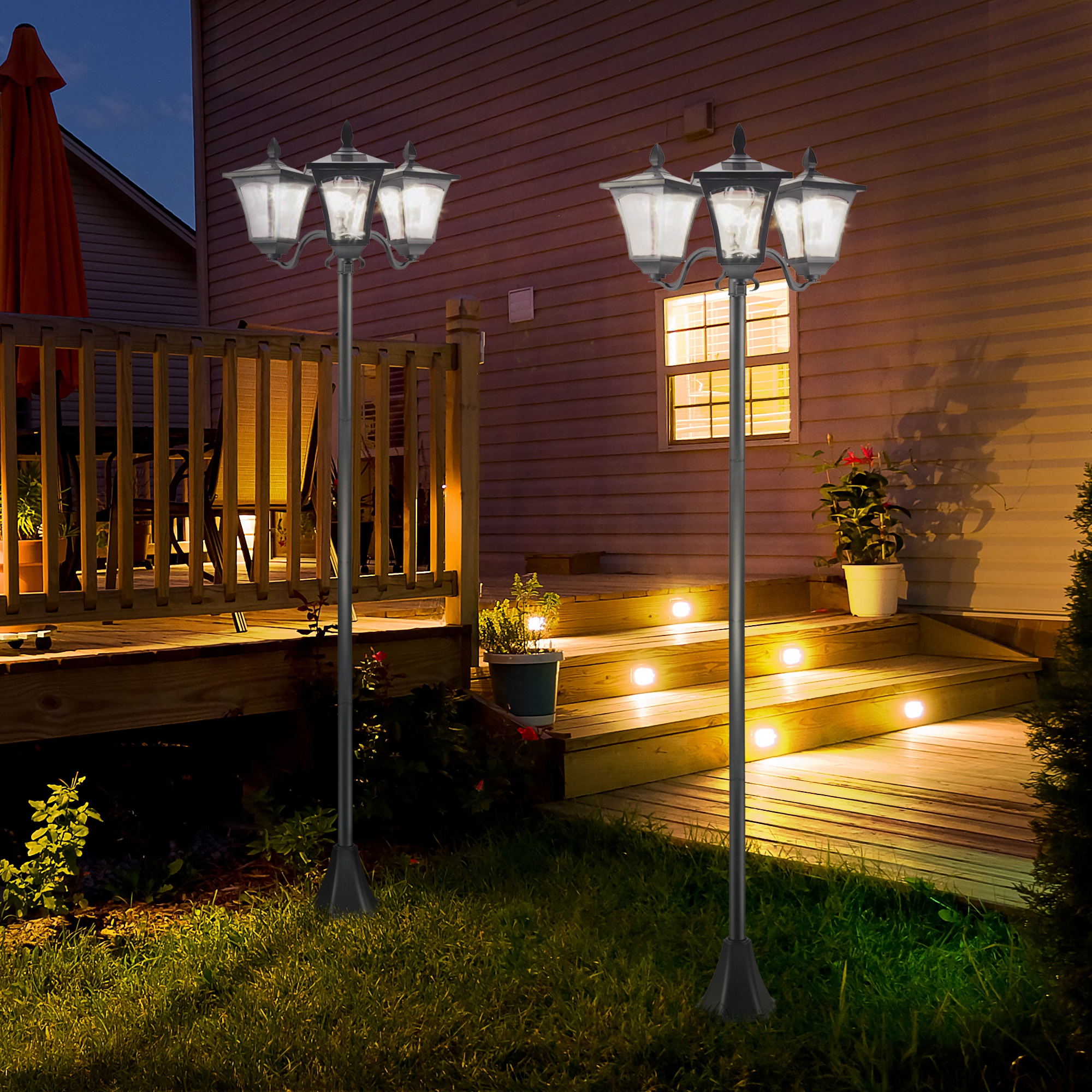 Dos farolas negras con múltiples luces están en un jardín por la noche. Una terraza de madera y una casa están en el fondo.
