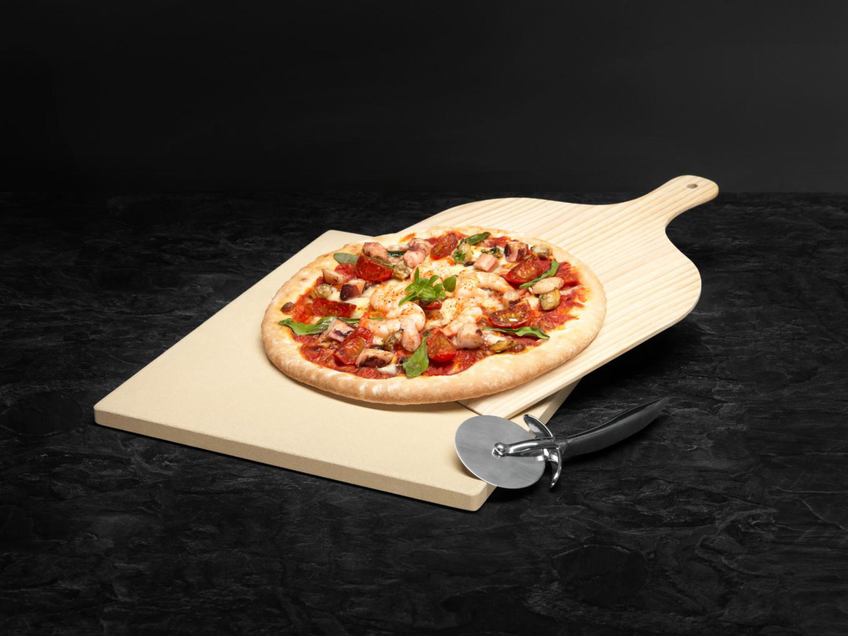 Una pizza en una tabla de madera y losa de piedra con un cortador de pizza sobre una superficie negra.
