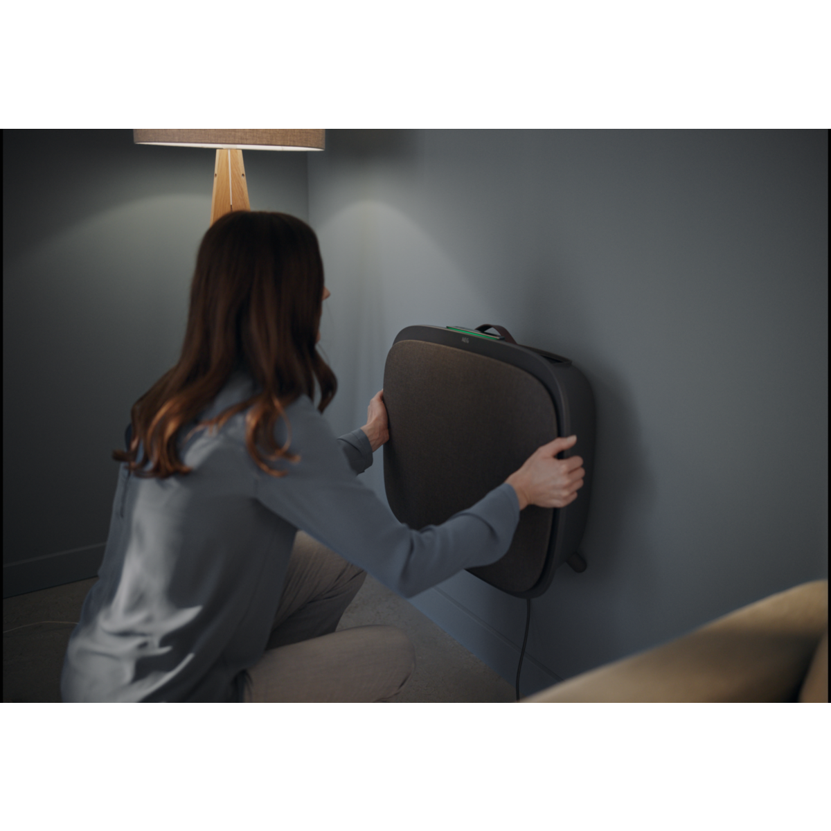 Una mujer colocando un purificador de aire negro en una pared gris. Una lámpara y un sofá son visibles en el fondo.