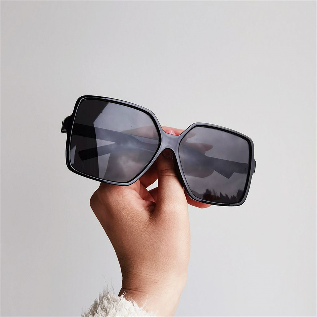 Die Hand einer Person, die eine schwarze Sonnenbrille hält. Der Hintergrund ist eine helle Wand.