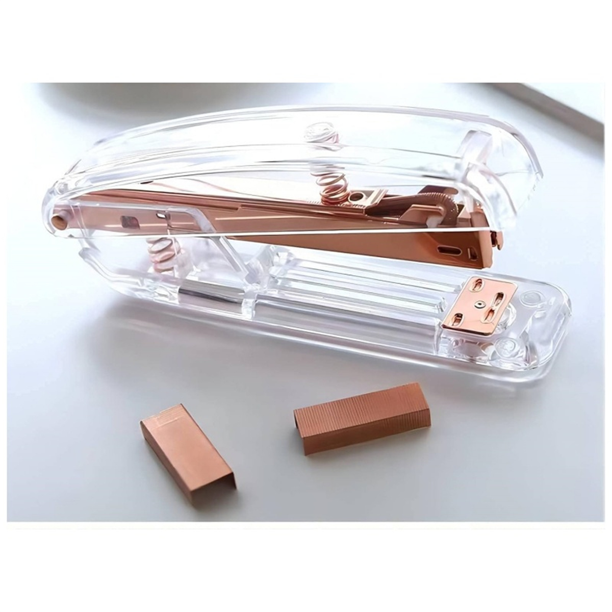 Ein klarer Tacker mit roségoldenen Innenteilen und zwei roségoldenen Heftklammern auf weißer Oberfläche.