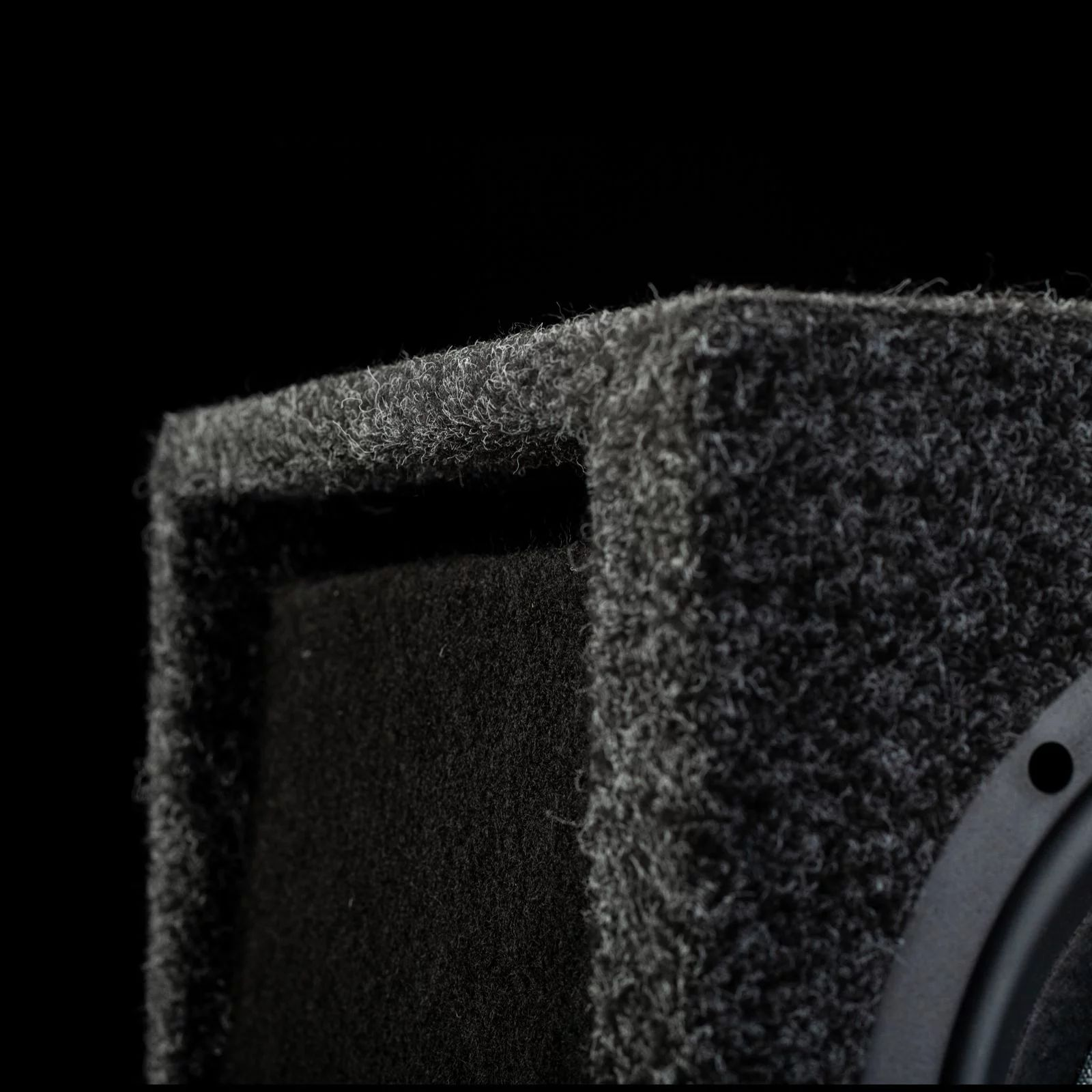 Nahaufnahme eines Subwoofer-Gehäuses. Schwarze Oberfläche, strukturiertes Material, dunkler Hintergrund.