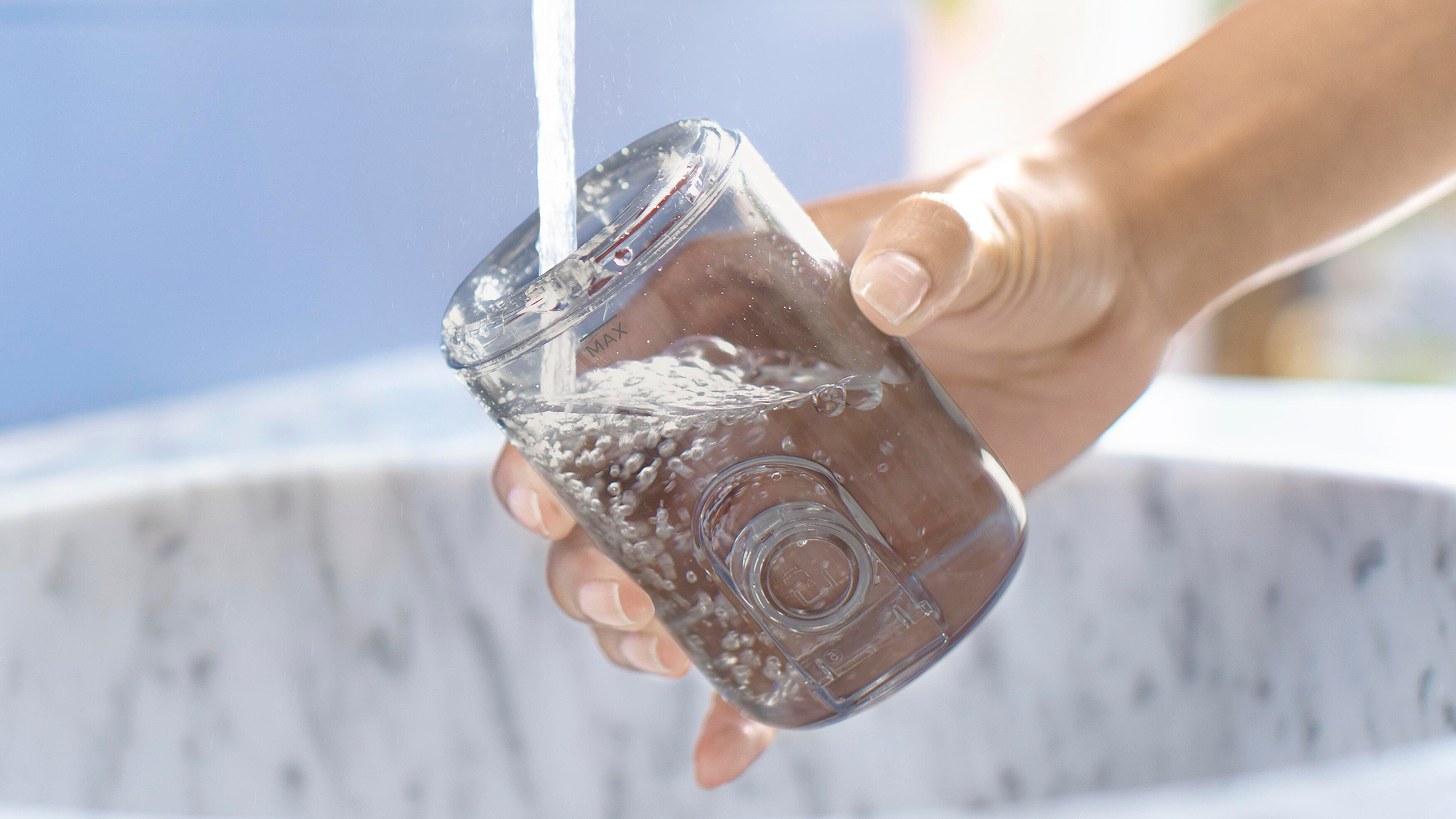 Hand hält transparenten Behälter, Wasser gießt hinein, Blasen sichtbar, neben einem Marmorwaschbecken.
