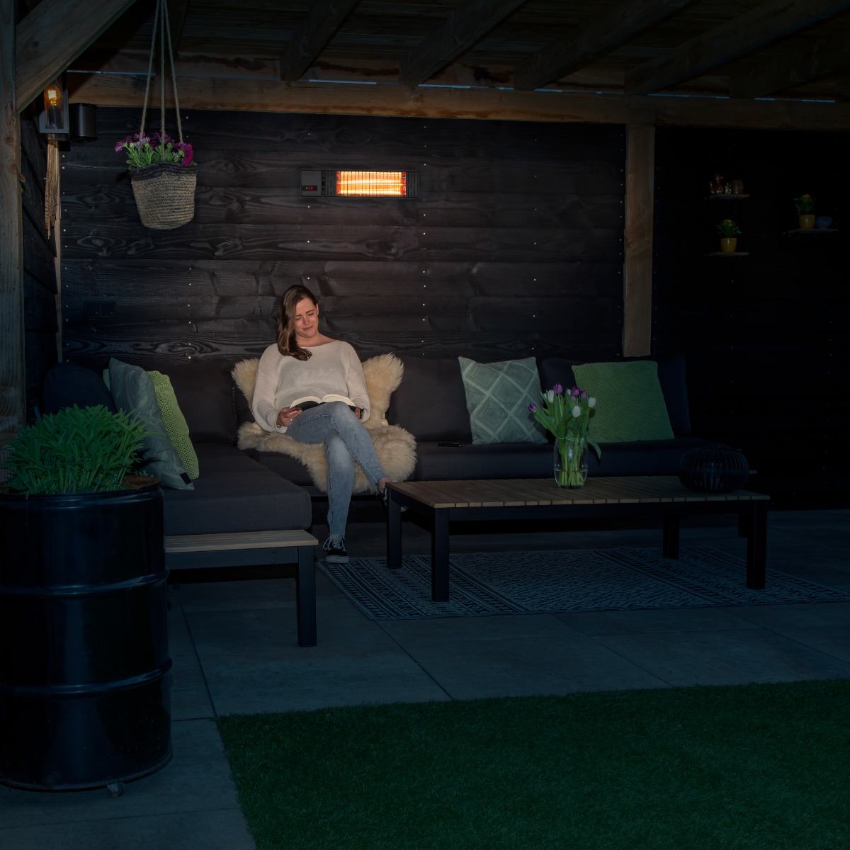 A woman reading under a covered patio with a heater, plants, and a table.